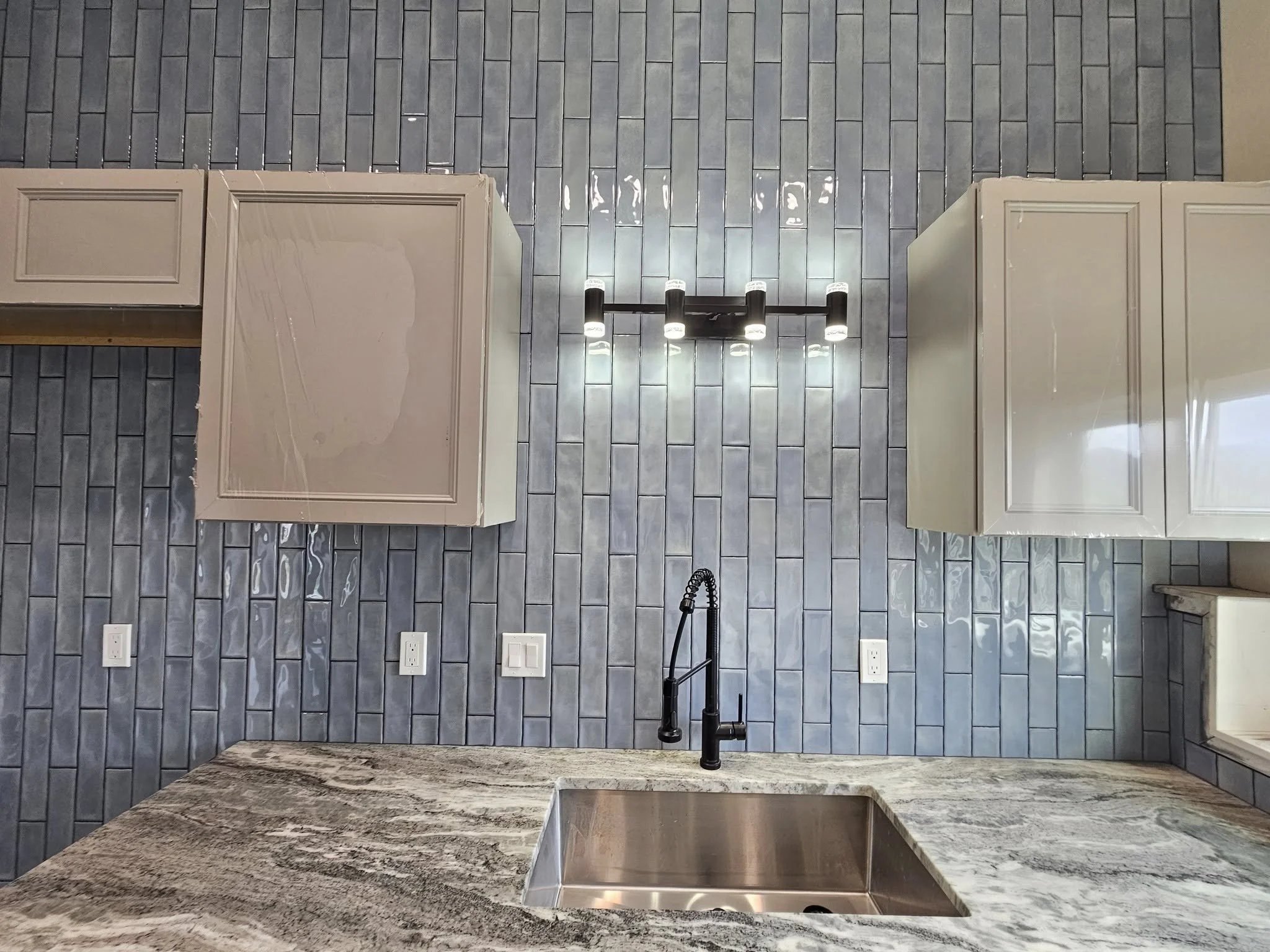 Overhead view of a kitchen counter with a sink, black faucet, and gray cabinets, with a dark gray tile backsplash and a wall-mounted light fixture.