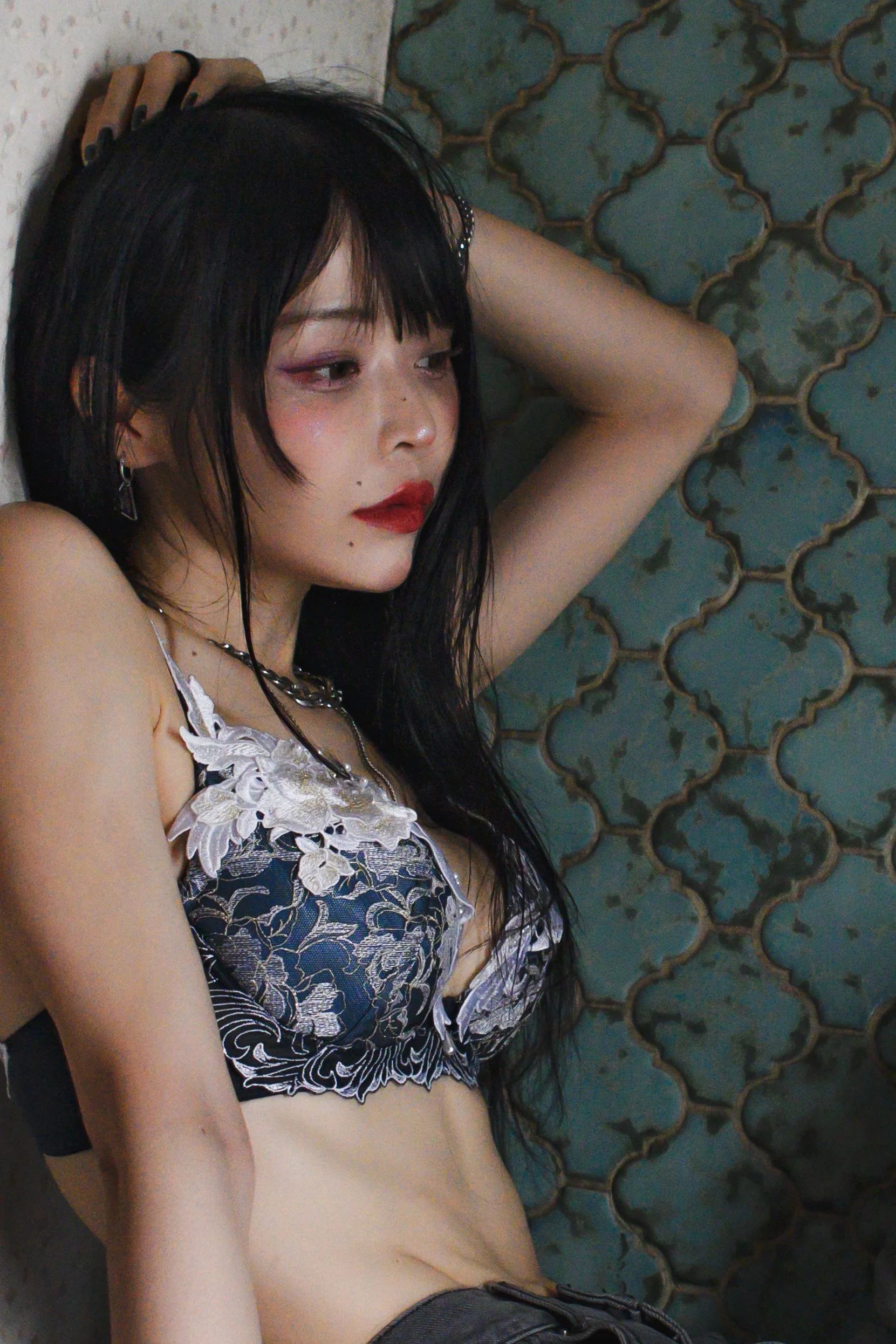 A young woman with black hair lying on the floor, eyes closed, wearing a black and white floral lace bralette and jewelry, with a patterned blue and brown tiled wall in the background.