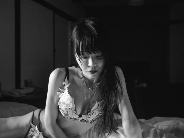 A woman with dark hair and bangs, wearing a lace bralette and layered necklaces, sitting on a bed in a dimly lit room.