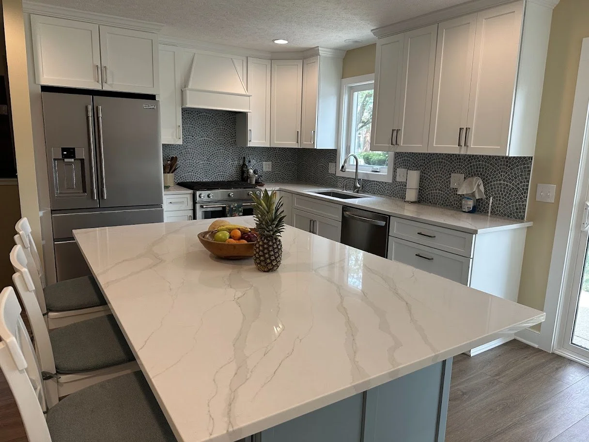Modern kitchen with white cabinets, gray backsplash, stainless steel refrigerator and appliances, a kitchen island with a pineapple and fruit bowl, and a window above the sink.