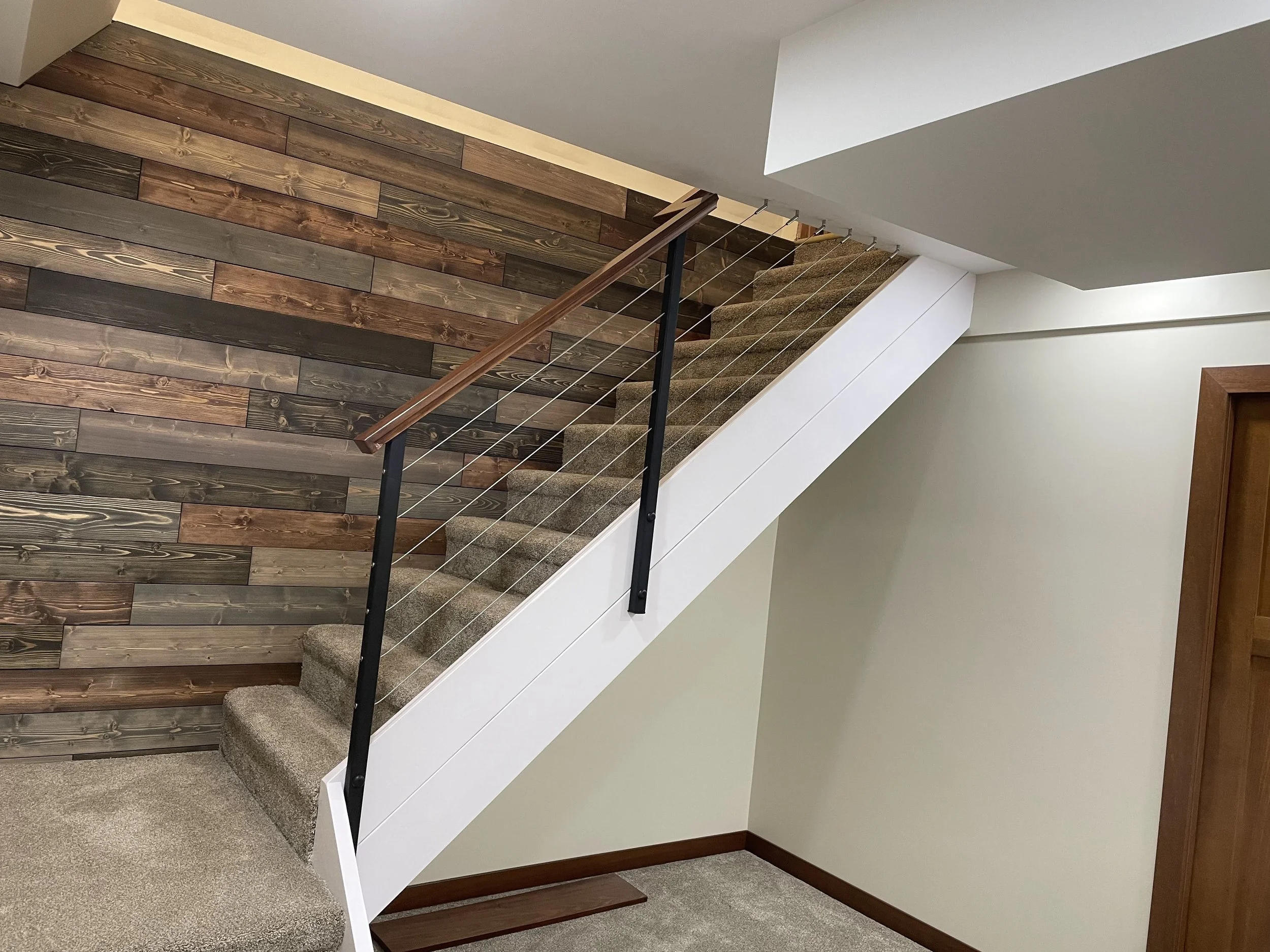 Interior view of a staircase with a wooden handrail, black metal supports, and wire balusters; the wall beside the staircase features wood paneling, and the surrounding walls are painted white.