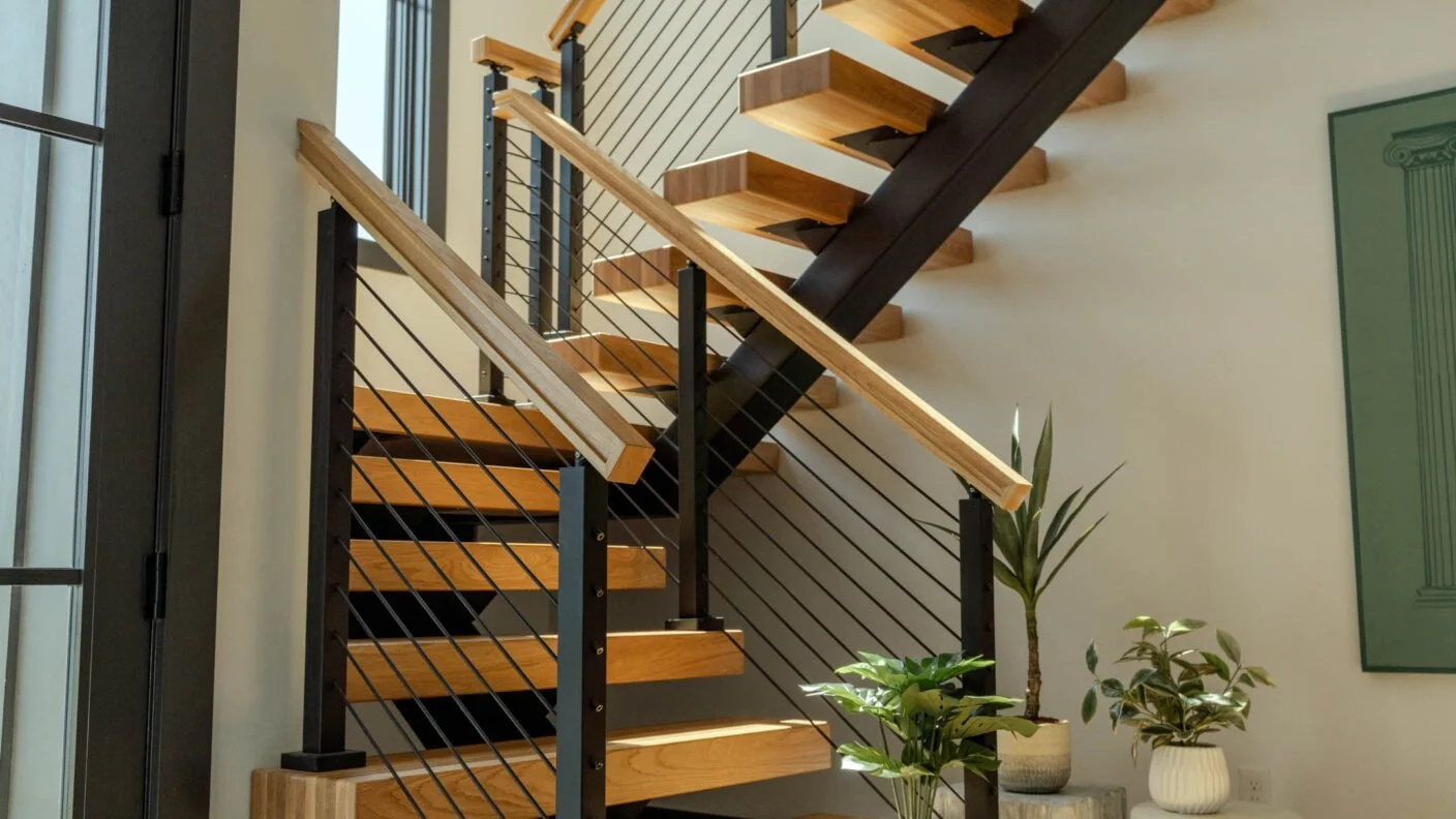 Interior staircase with wooden steps, black metal support, and cable railing, next to potted plants and a green wall art piece.