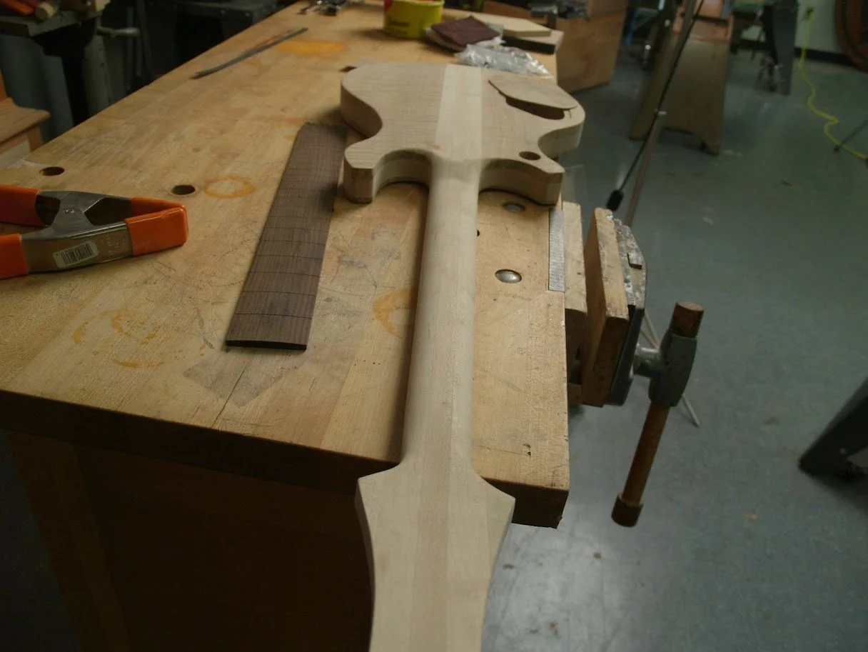 Unfinished electric guitar body on a workbench with a ruler and clamp nearby in a woodworking shop.