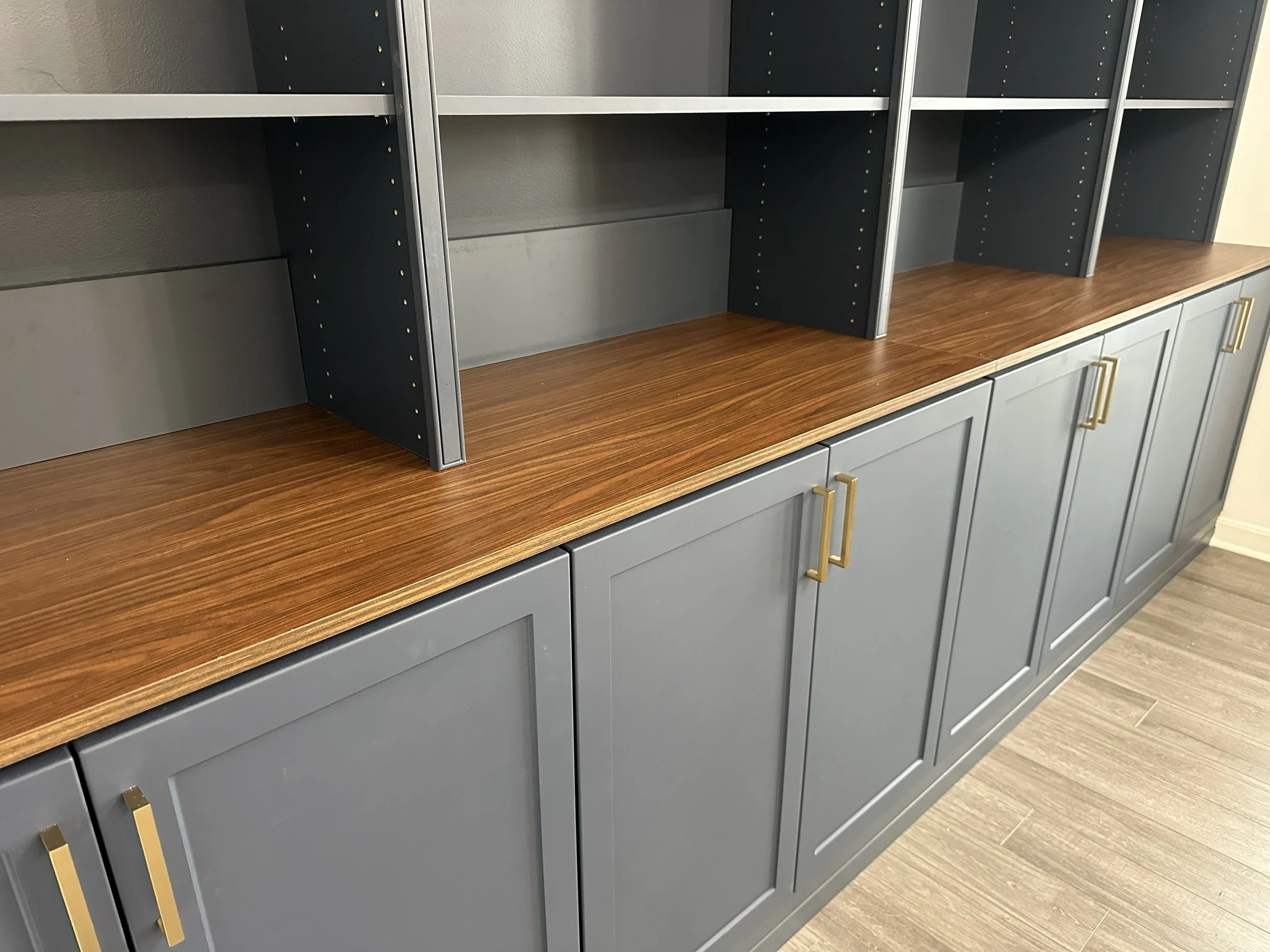 Grey cabinetry with gold handles and empty black shelves on top, wooden countertop, and wood-look flooring.
