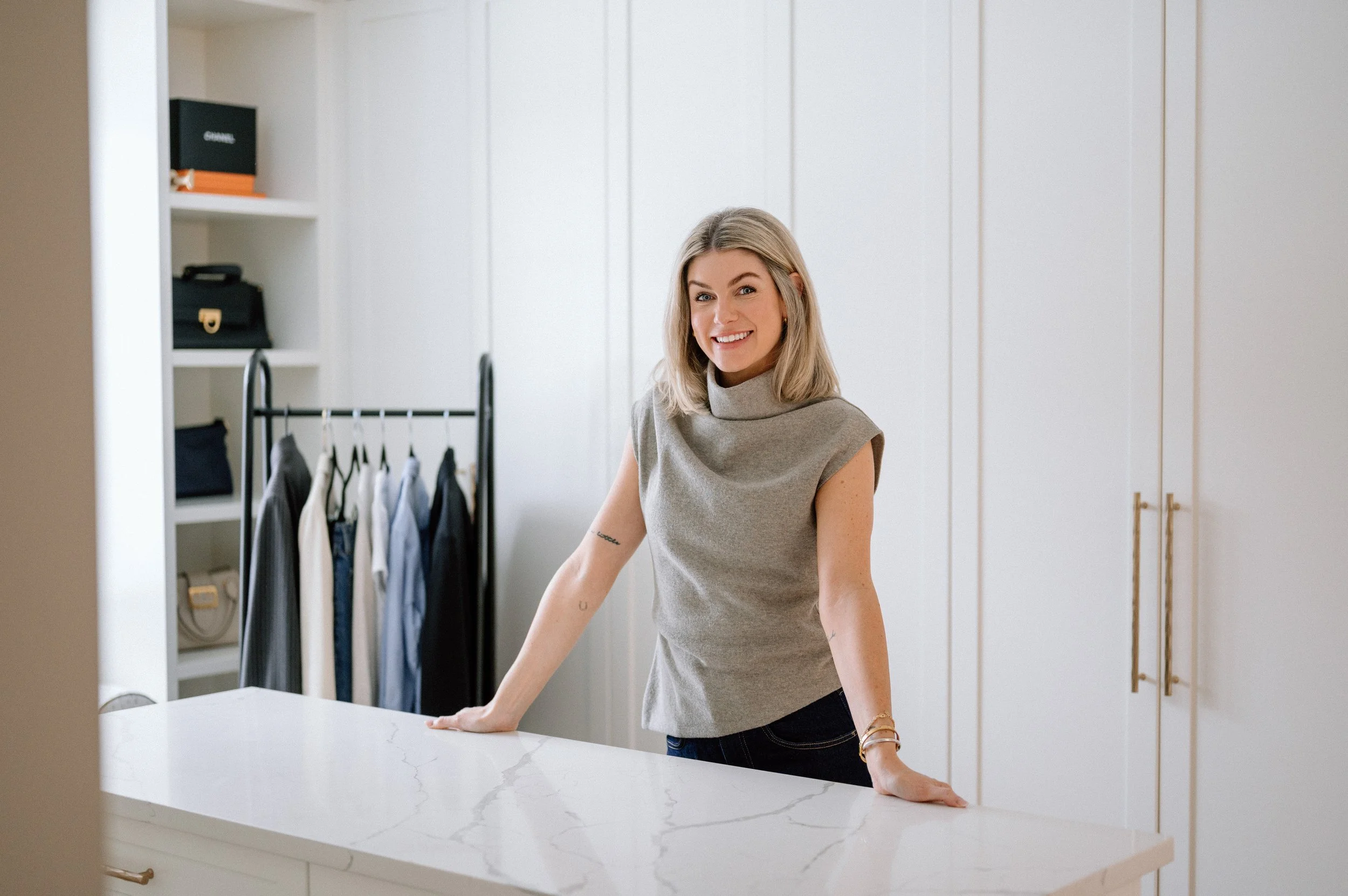 A woman with blonde hair smiling while leaning on a white marble kitchen island in a modern, white kitchen with closed cabinets and a clothing rack with hanging clothes in the background.