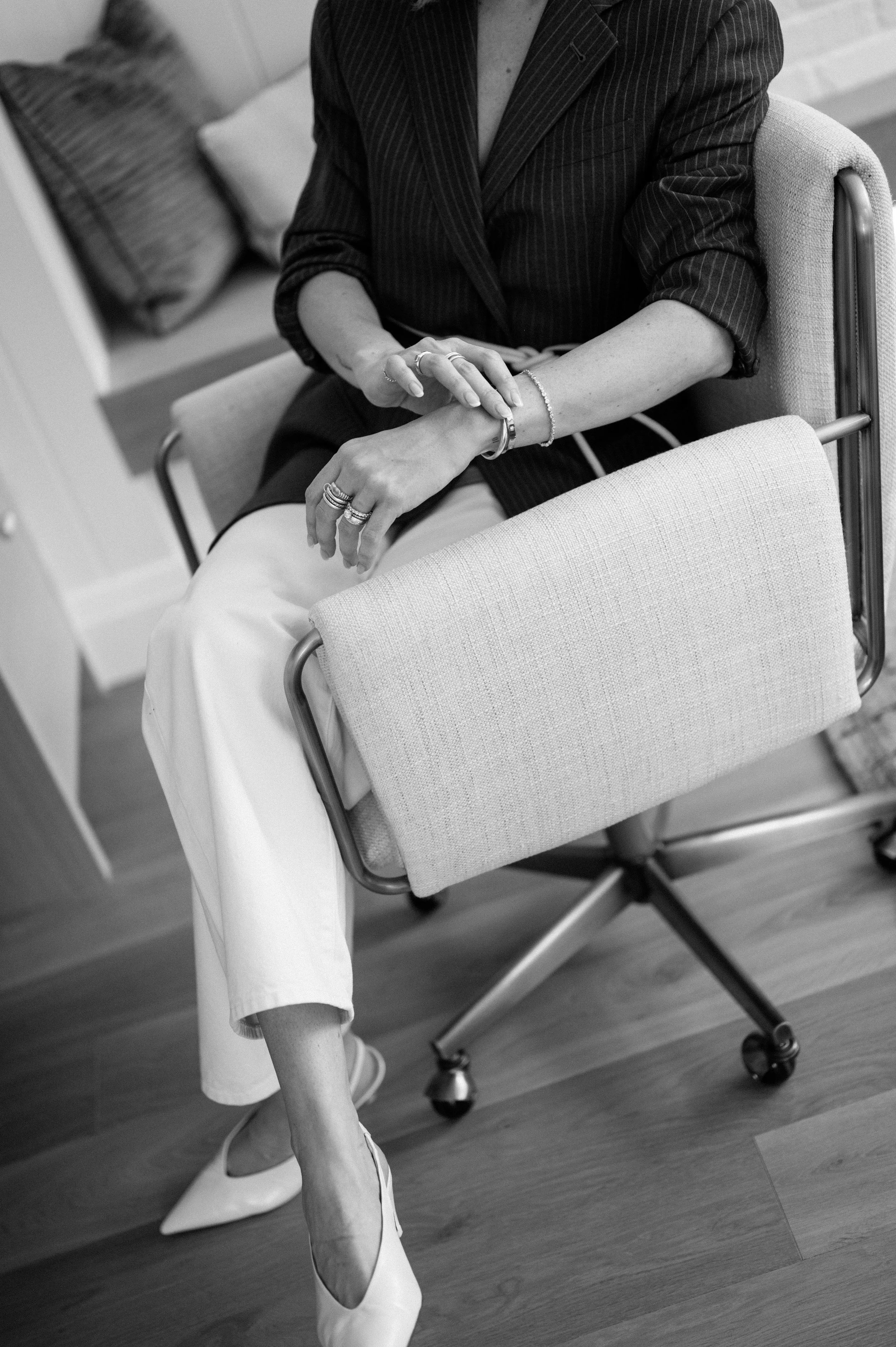 A woman sitting on an office chair, wearing a pinstripe blazer, white pants, and pointed-toe heels, with jewelry on her hands and wrists, in a room with a sofa and pillows in the background.