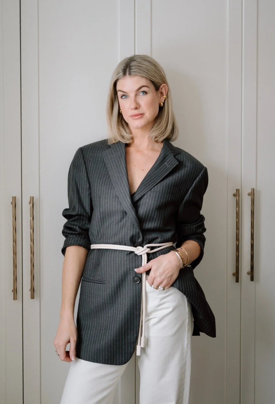 A woman with blonde hair, wearing a pinstripe blazer dress tied with a white belt, standing in front of white cabinets.