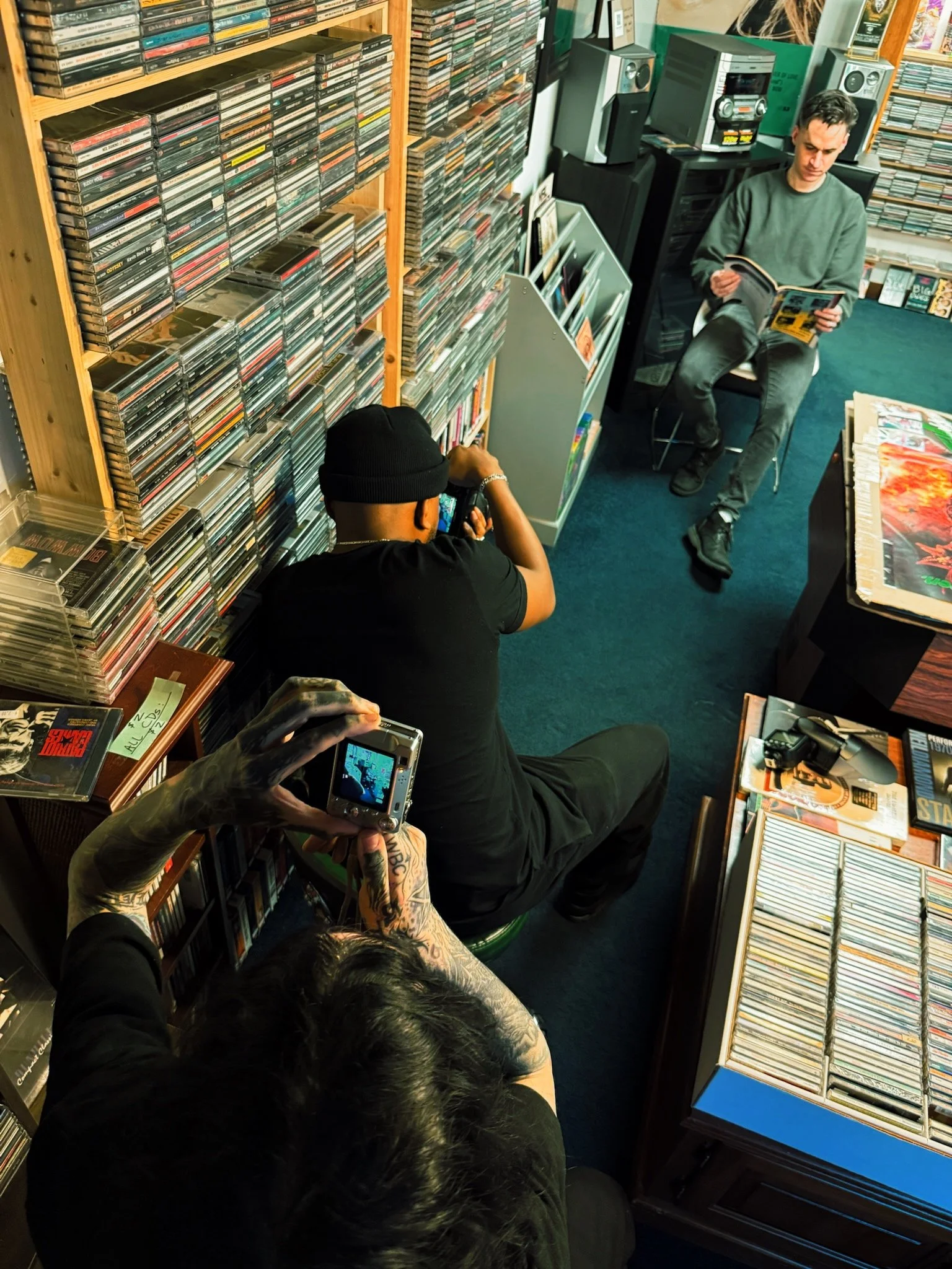 A record store with shelves full of CDs and vinyl records. A man with tattoos is taking a photo with a small digital camera, while a person in a black beanie is sitting and looking at his phone. In the background, a young man is sitting on a chair reading a comic book. The store has a carpeted floor and various music-related items on display.