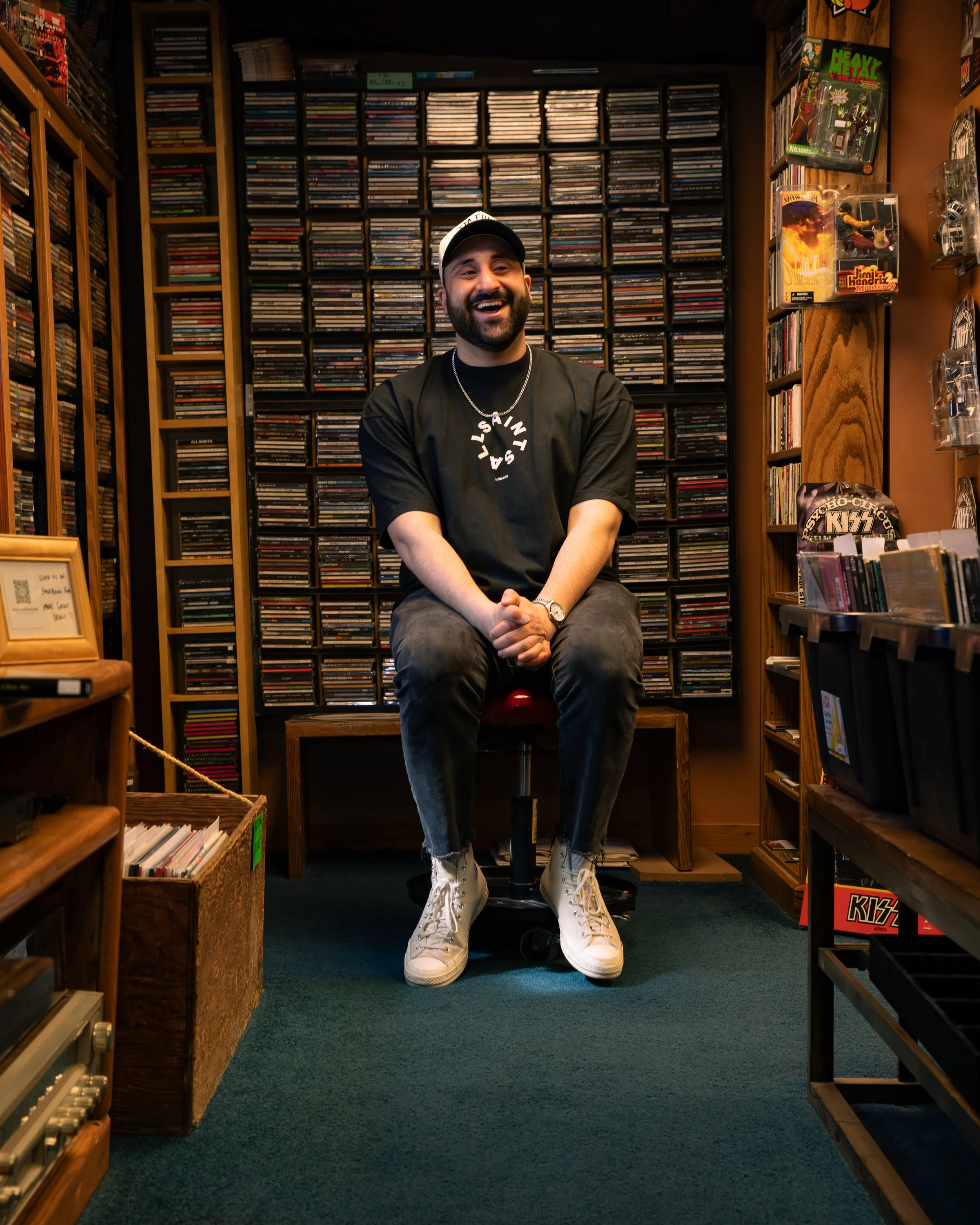 A man with a beard wearing a black T-shirt that says 'Fear of God Essentials' and a black cap, standing behind DJ equipment with colorful lights in a nightclub or bar setting.