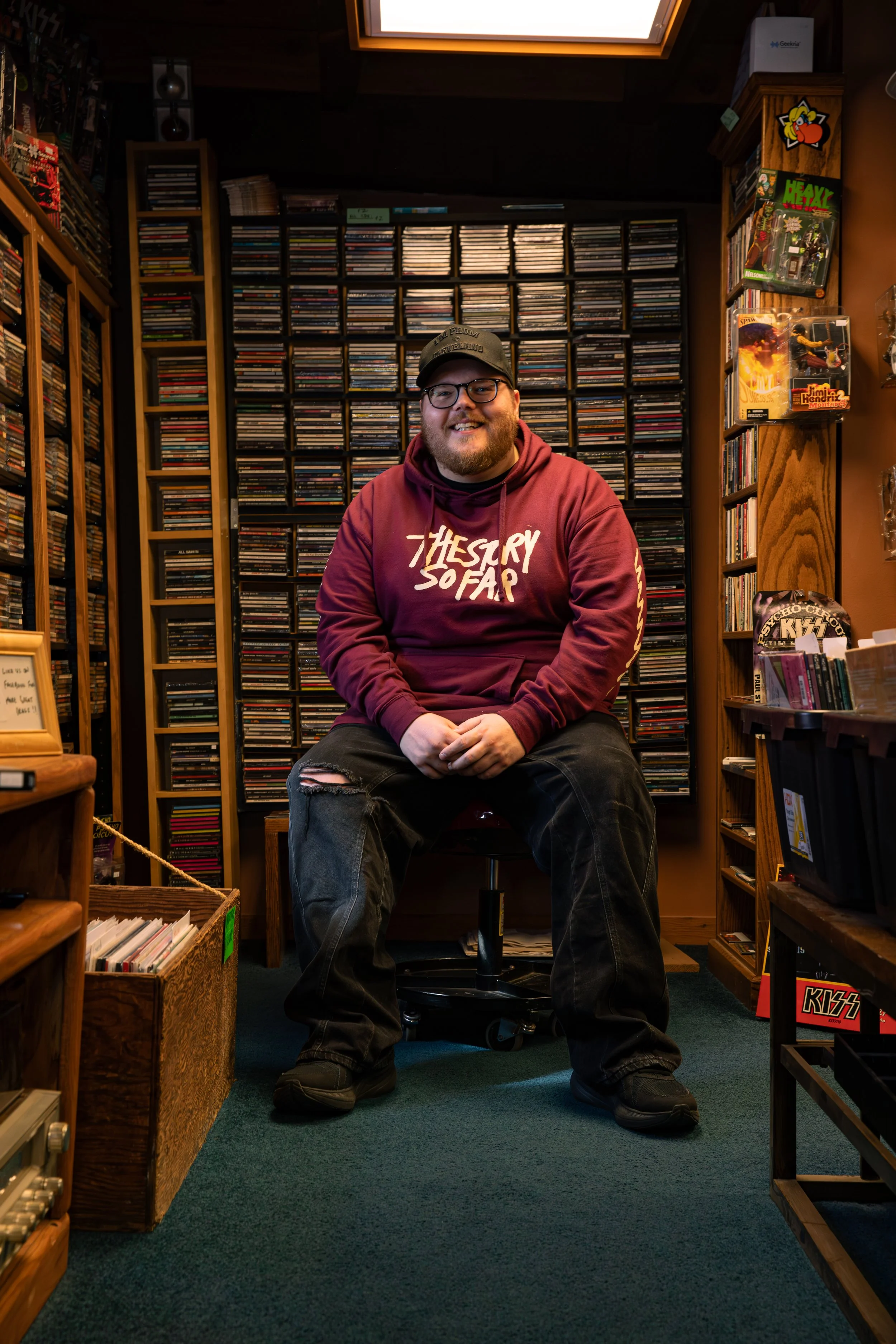 A smiling man with glasses and a beard, wearing a black shirt, resting his chin on his hand, showcasing colorful tattoos on his arms, against a neutral gray background.