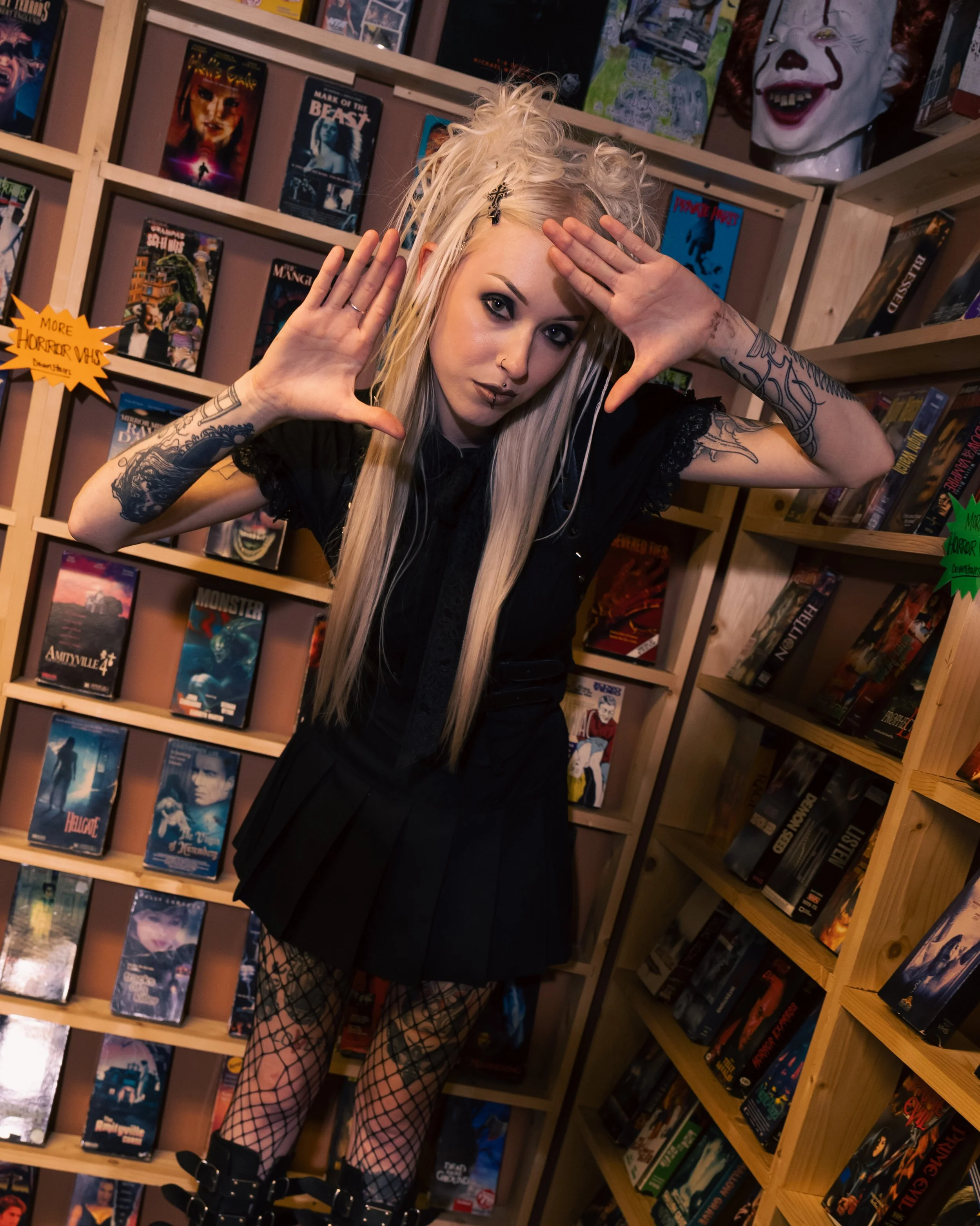 Two women with goth style inside a store that sells horror movies on shelves. The woman in front has long black hair, glasses, black lipstick, and is reaching towards the camera. The woman in the background has long blonde hair, and a confident pose leaning on the shelf.