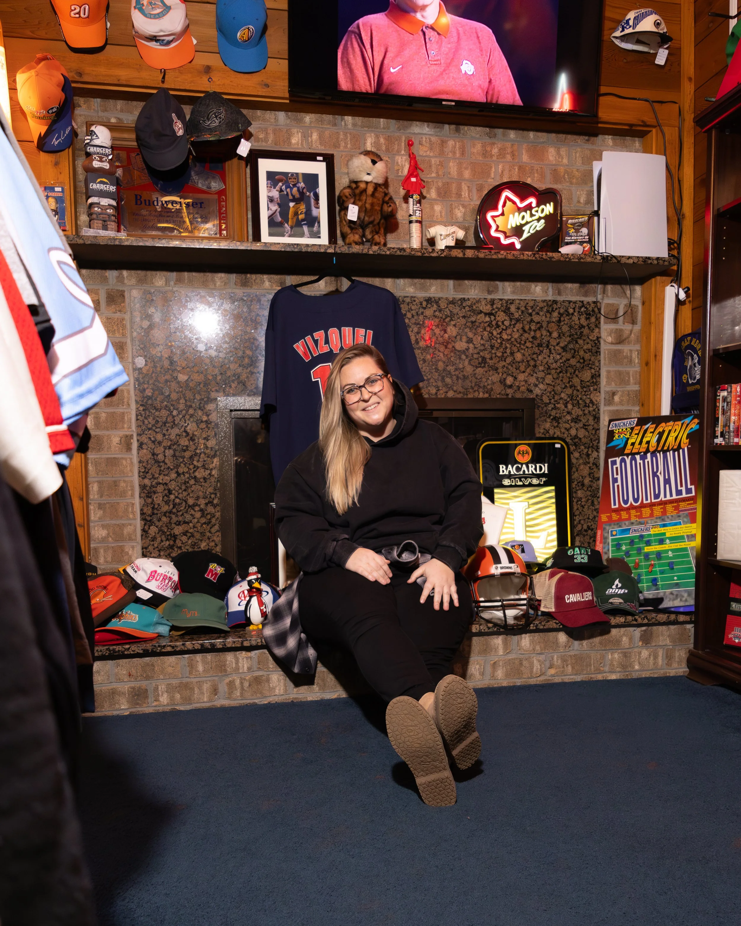 Close-up of a smiling woman with glasses and curly hair, wearing hoop earrings, layered necklaces, and a striped shirt, in an indoor setting.
