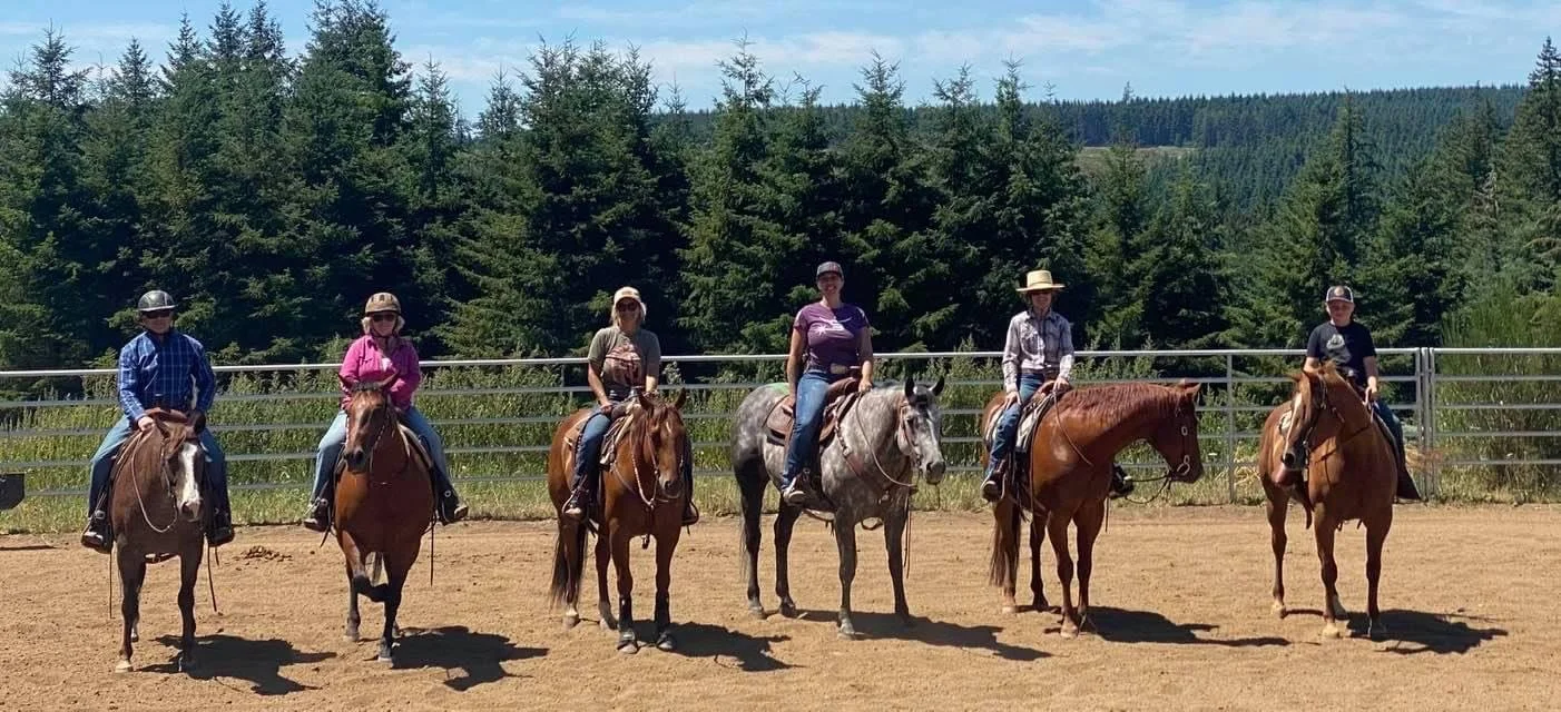 Six people riding horses in a fenced outdoor riding arena with trees and hills in the background.