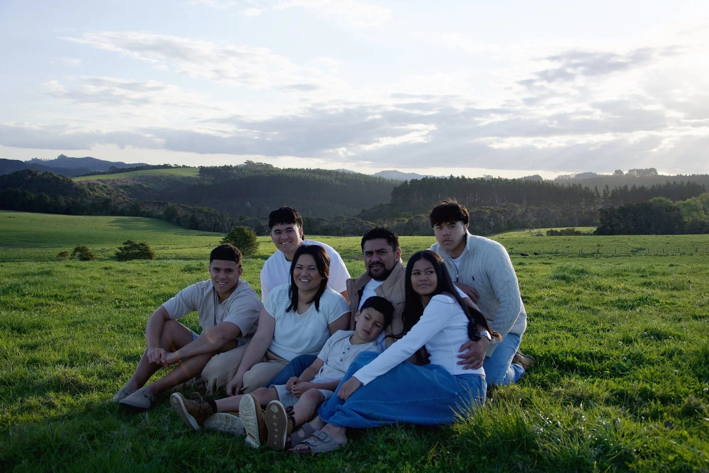 A group of people sitting on grass in a scenic outdoor setting with rolling hills and trees in the background under a cloudy sky.