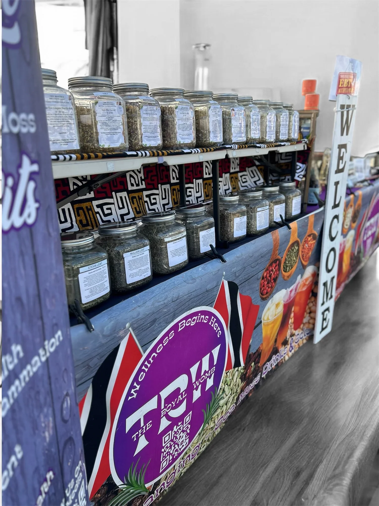 Display of glass jars filled with herbs and spices on a table at a market stall, with colorful signage and promotional posters in the background.