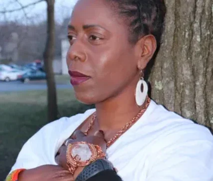 A woman leaning against a tree, wearing earrings, a watch, and a white shirt, with a solemn expression.