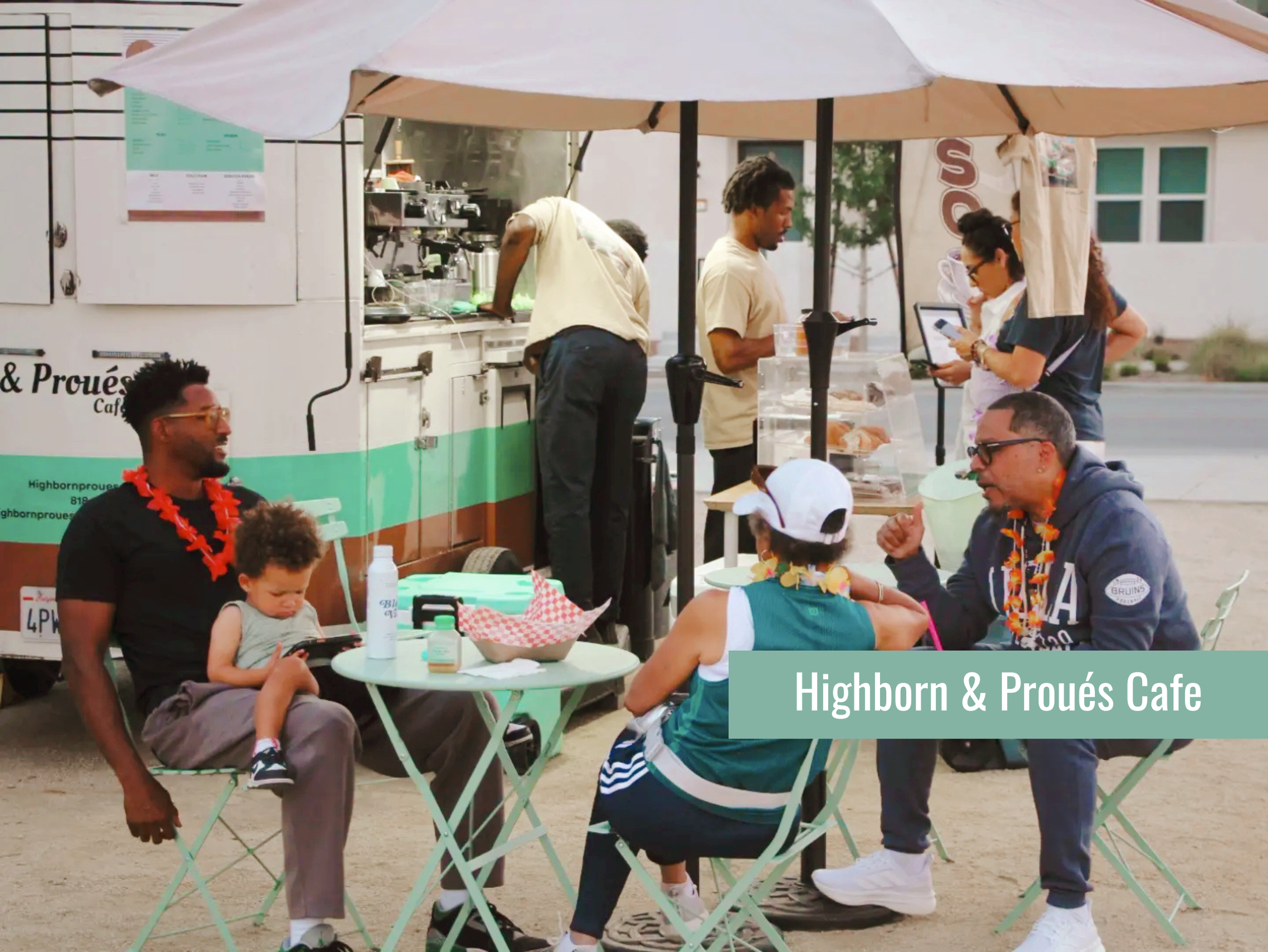 People enjoying food and socializing at an outdoor cafe stand called Highborn & Prouès Cafe, with a man and a woman standing near the counter, and others sitting at tables under umbrellas.