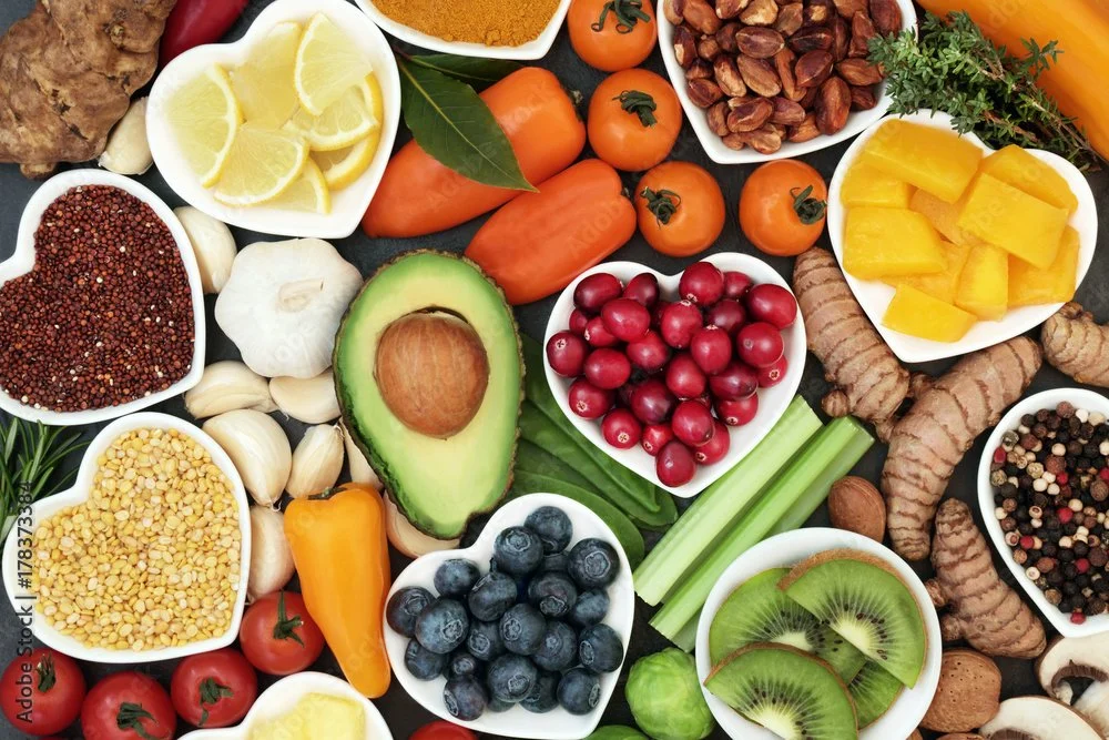 Assorted fresh fruits and vegetables, including cherry tomatoes, blueberries, kiwi slices, avocado, lemon wedges, pineapple chunks, grapes, garlic, turmeric roots, blackberries, yellow lentils, pink peppercorns, and various herbs.
