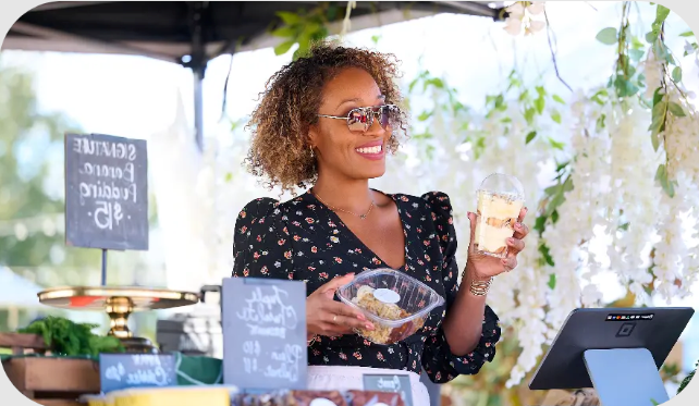A woman with curly hair, sunglasses, and a floral dress smiling and holding a container of food and a sandwich at an outdoor market or festival, with a digital payment device in front of her and blurred greenery in the background.