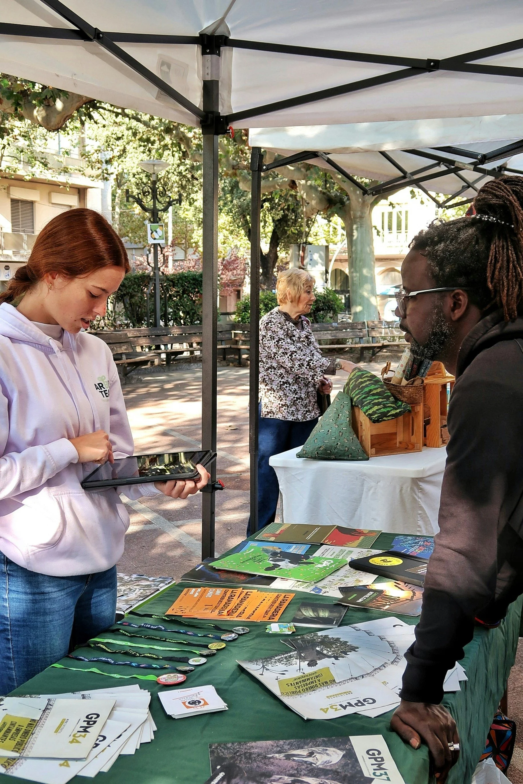 A park scene with a booth or table displaying various brochures, buttons, and flyers related to wildlife or environmental conservation. A woman in a white hoodie is using a tablet, and a man with glasses is leaning on the table. In the background, an older woman is looking at items on another table, with trees and park benches visible.