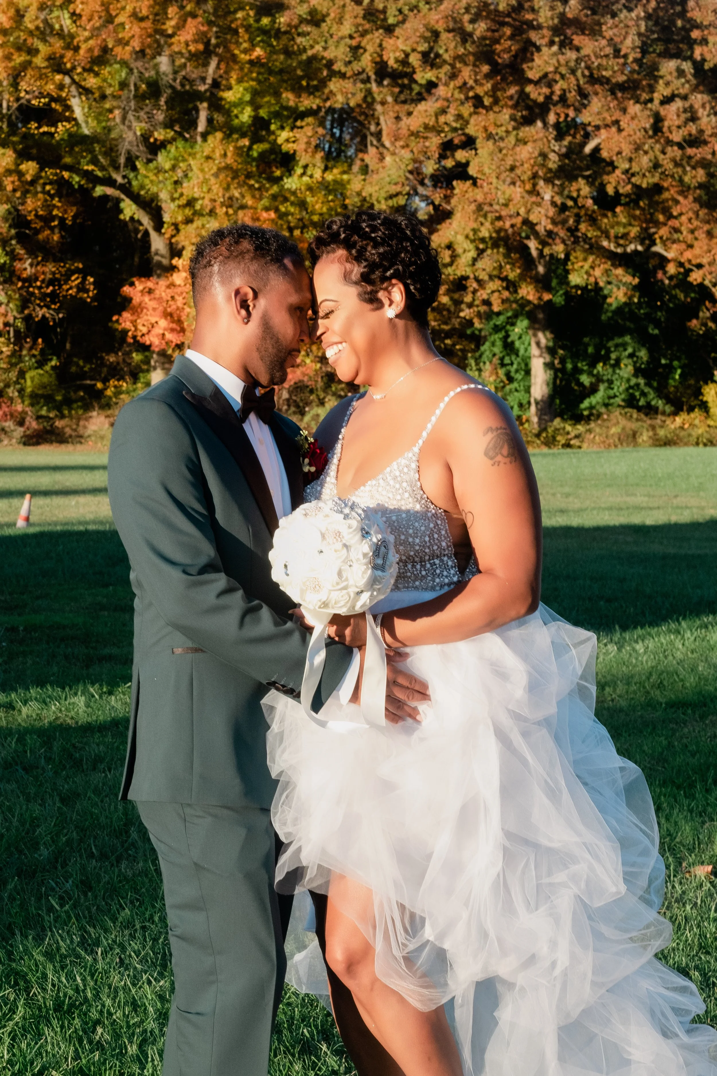 A bride and groom embrace outdoors in a park with colorful fall trees in the background, both smiling, the bride holding a white bouquet, wearing a sparkly wedding dress and the groom in a black tuxedo.
