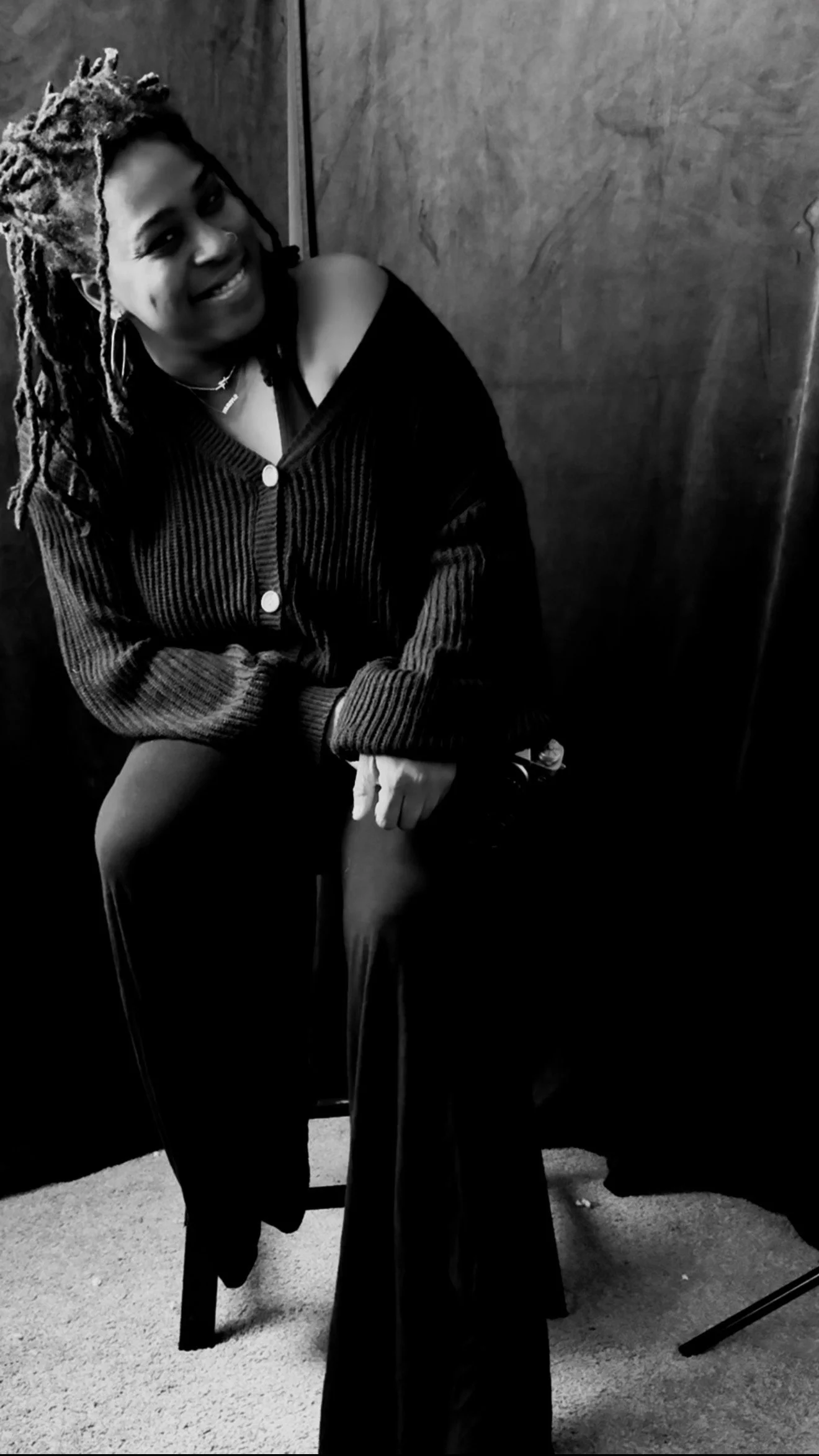 A woman with dreadlocks smiling and sitting on a stool in a room with wooden walls, wearing a dark off-the-shoulder sweater and dark pants, in black and white.