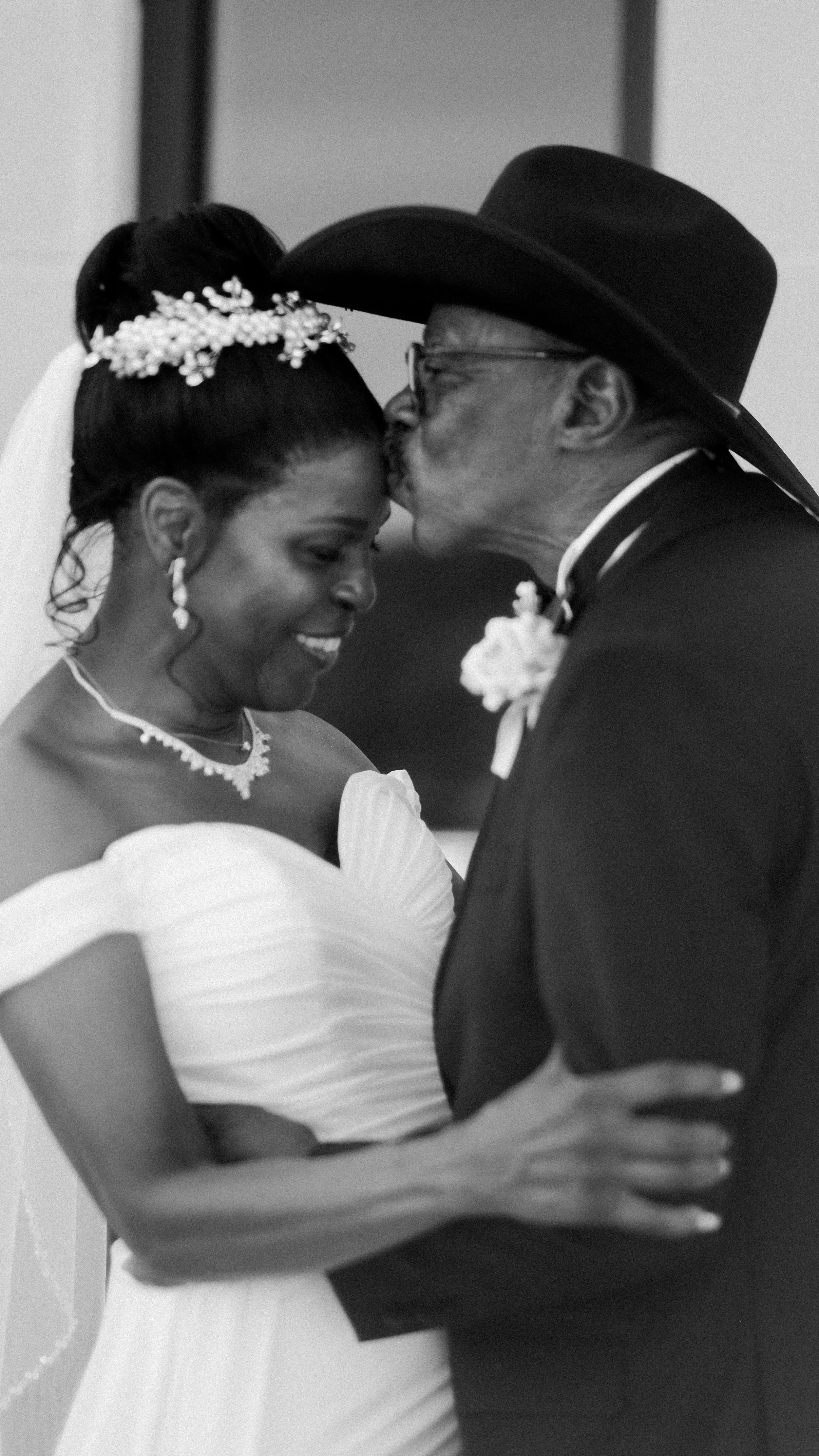 Black and white photo of a bride and an elderly man sharing a kiss, likely at her wedding.