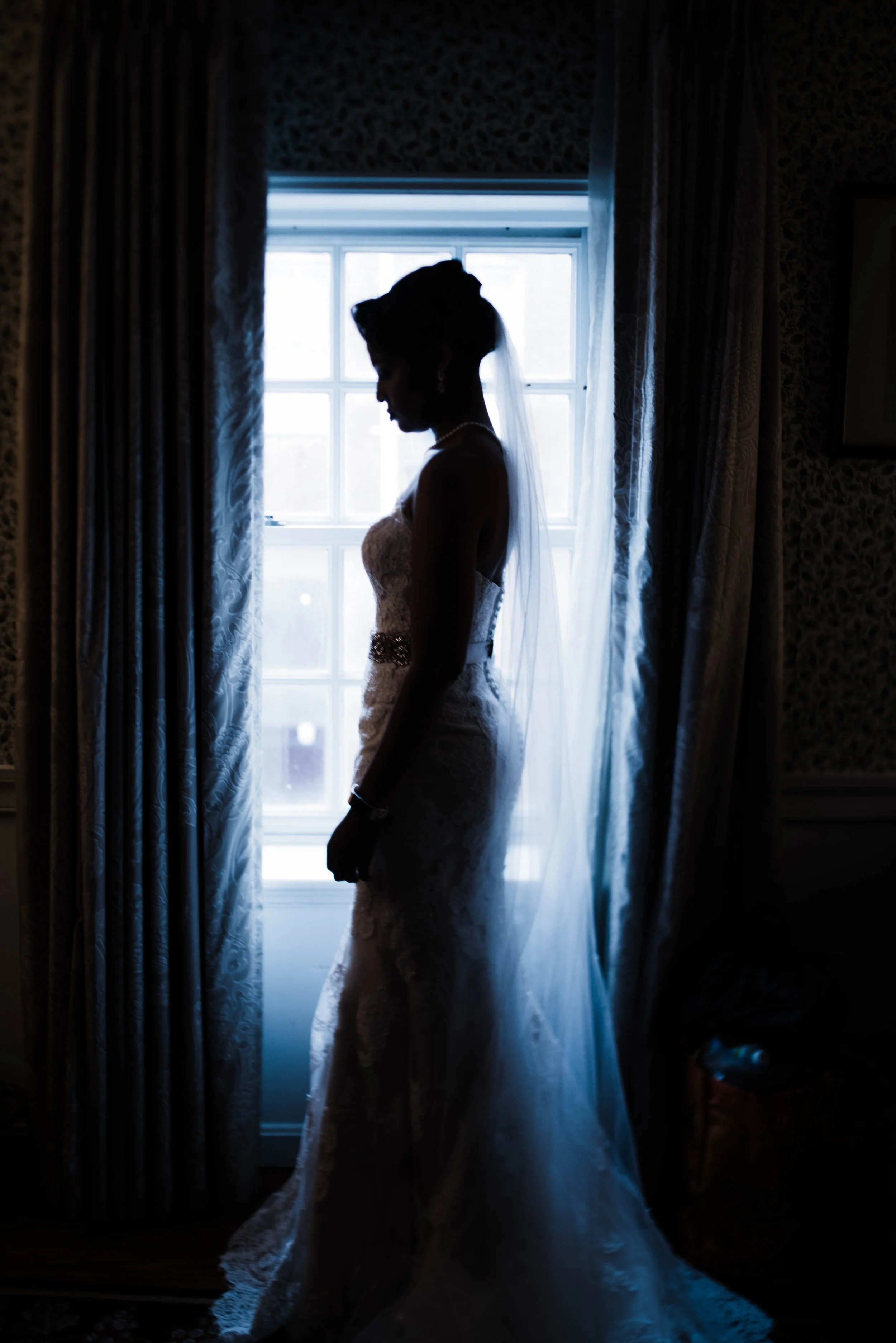 Silhouette of a bride in a wedding dress, standing by a window with curtains, backlit by natural light.