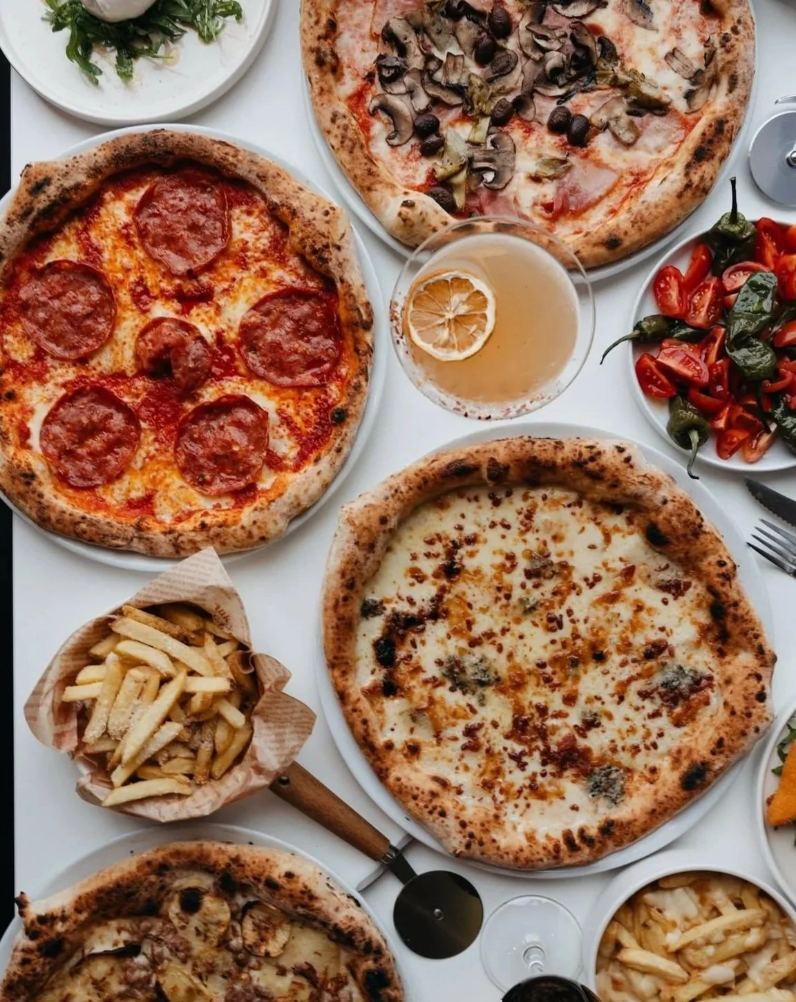 Several pizzas with various toppings, a bowl of French fries, a salad, sliced peppers, and a drink with a lemon slice.