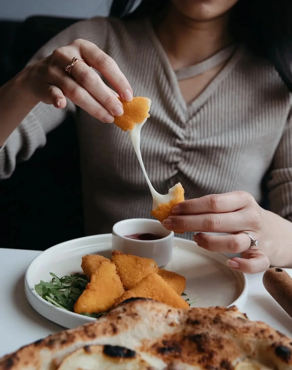 Woman dipping mozzarella sticks into marinara sauce at a restaurant table with pizza in the foreground.