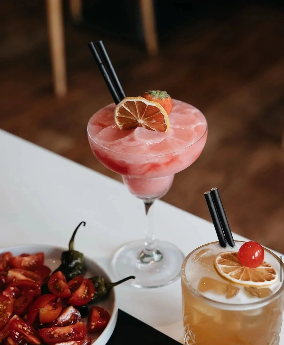 Two cocktails with dried citrus slices and cherries garnishes, alongside a bowl of cherry tomatoes and peppers.