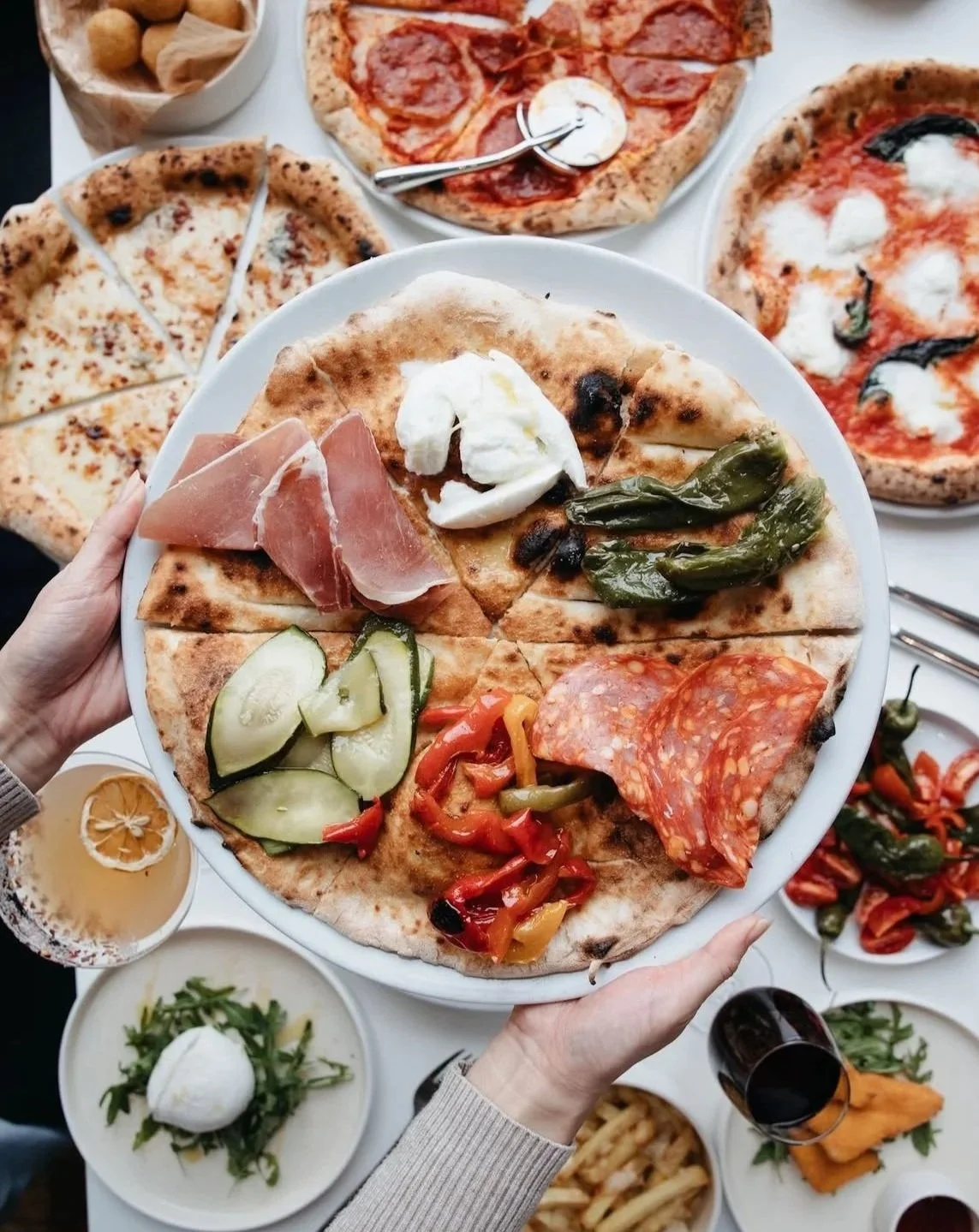 Plate of assorted pizza slices with toppings including mozzarella, ham, green peppers, cucumbers, and pepperoni, surrounded by additional pizza slices, food dishes, and drinks.