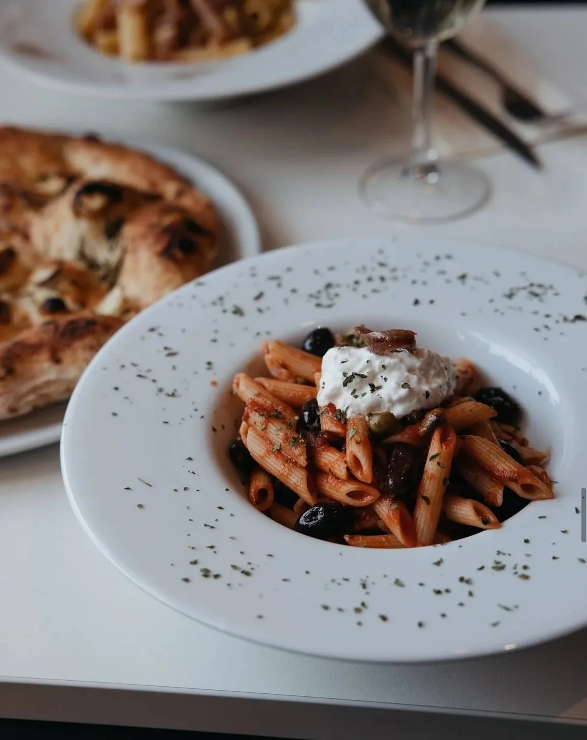 Plate of penne pasta with black olives and a topping of ricotta cheese, garnished with herbs, with a side of bread, on a table with a glass of wine in the background.