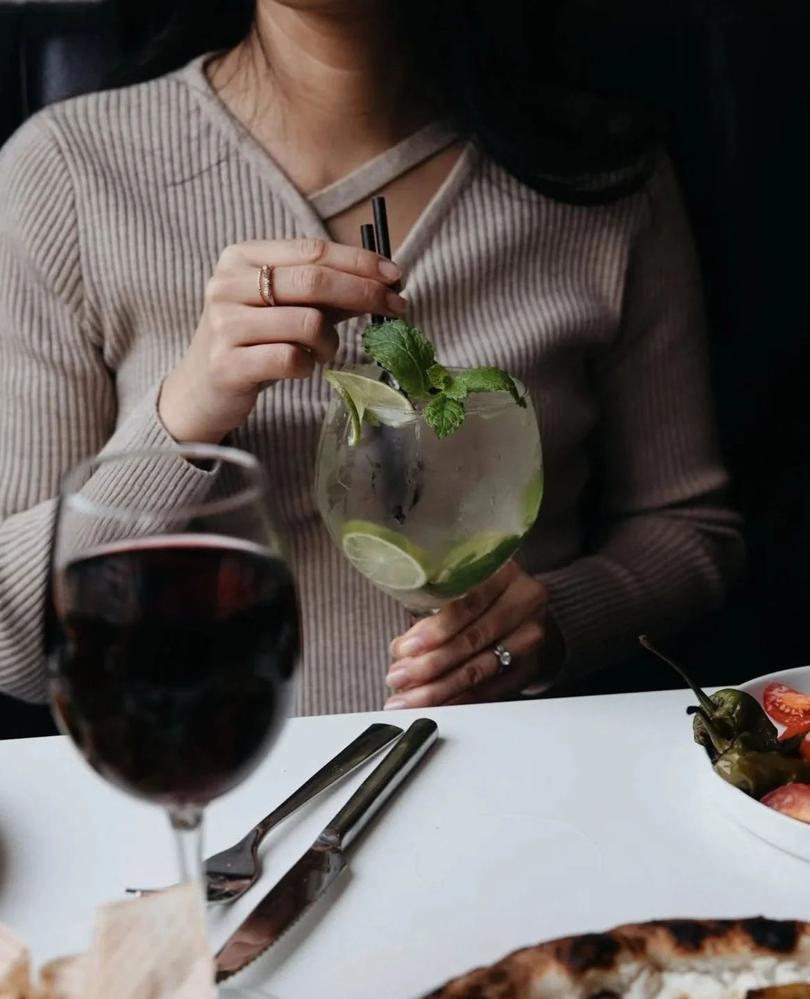 A woman holding a glass of mojito cocktail garnished with mint leaves, lemon slices, and limes at a table with a glass of red wine, cutlery, and a bowl of salad.
