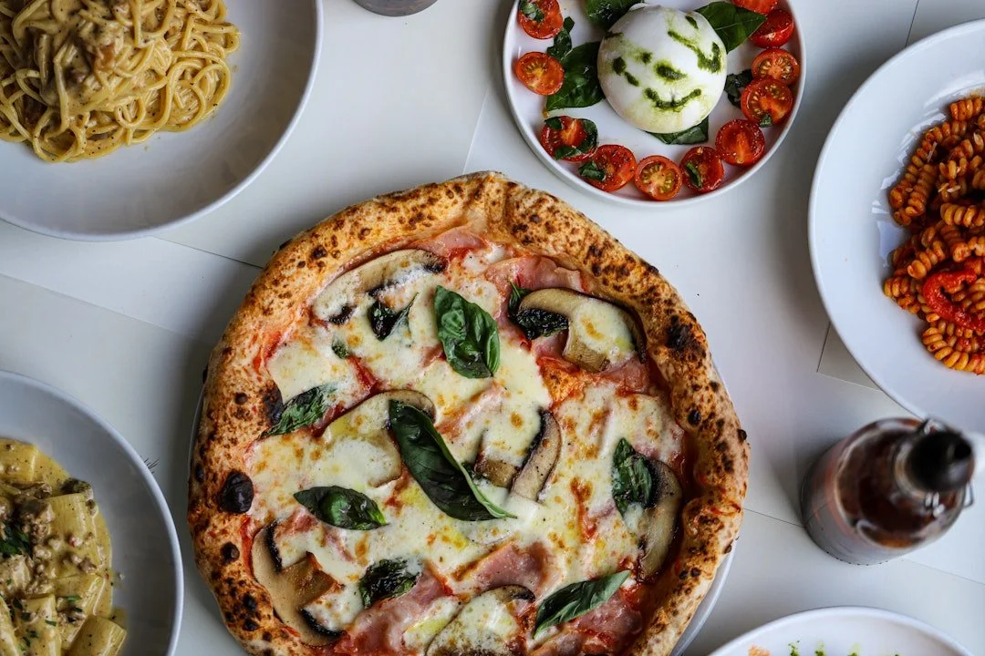 Slice of pizza with mushrooms, ham, and basil on a white table surrounded by plates of pasta, salad with tomatoes and a smiley face created with basil and tomato slices, and a bottle of soda.