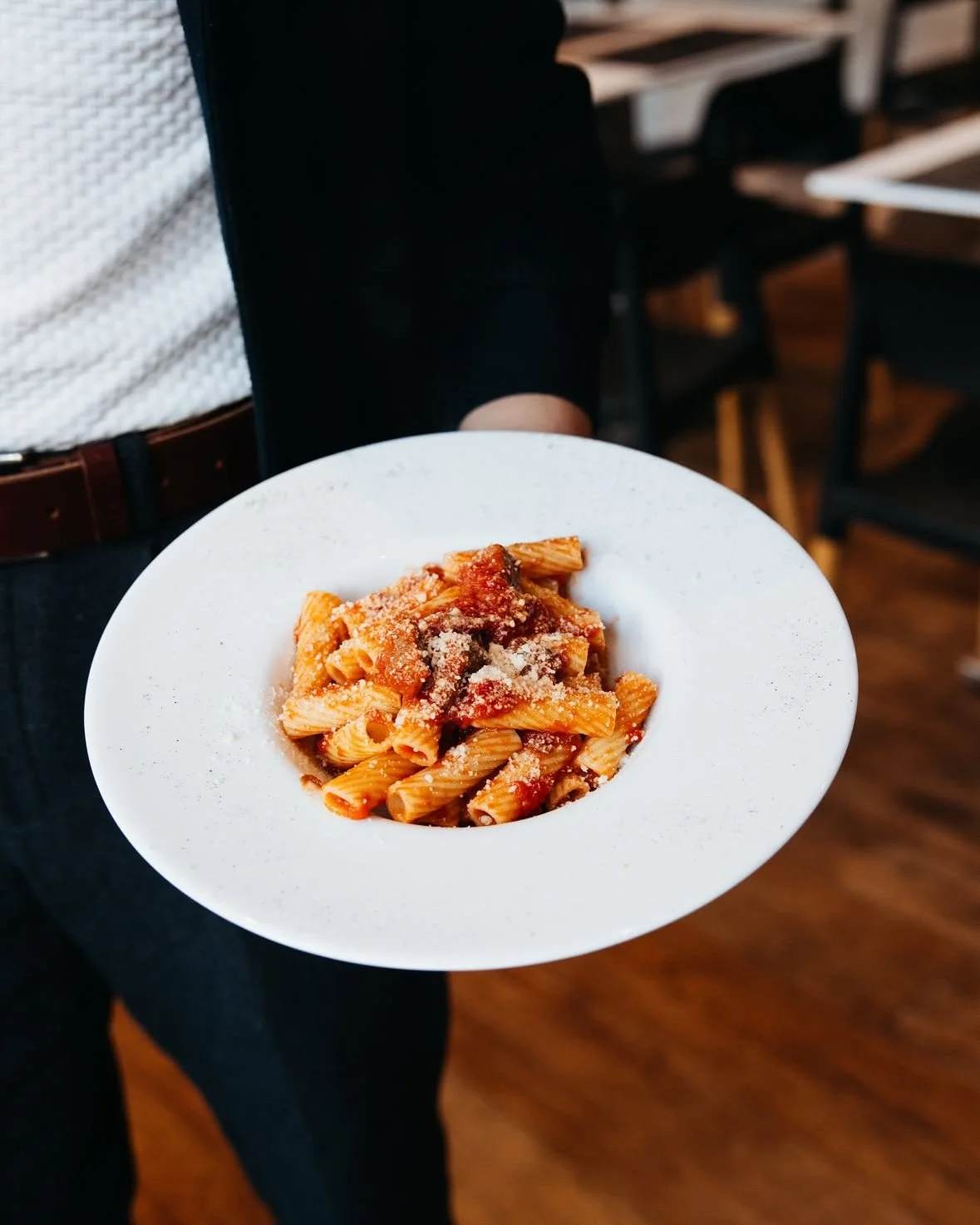 A person holding a white plate of cooked pasta with tomato sauce and grated cheese in a restaurant setting.