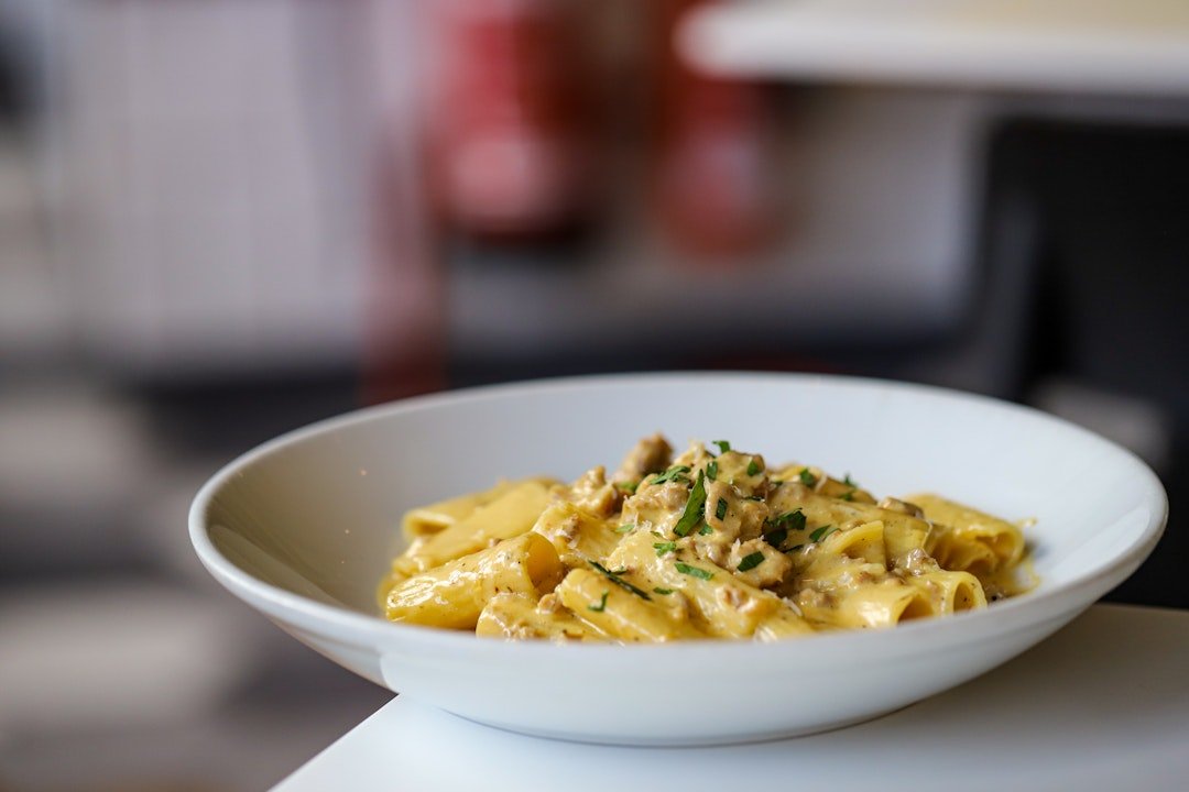 A white bowl of creamy pasta with chopped herbs on top, placed on a white surface.