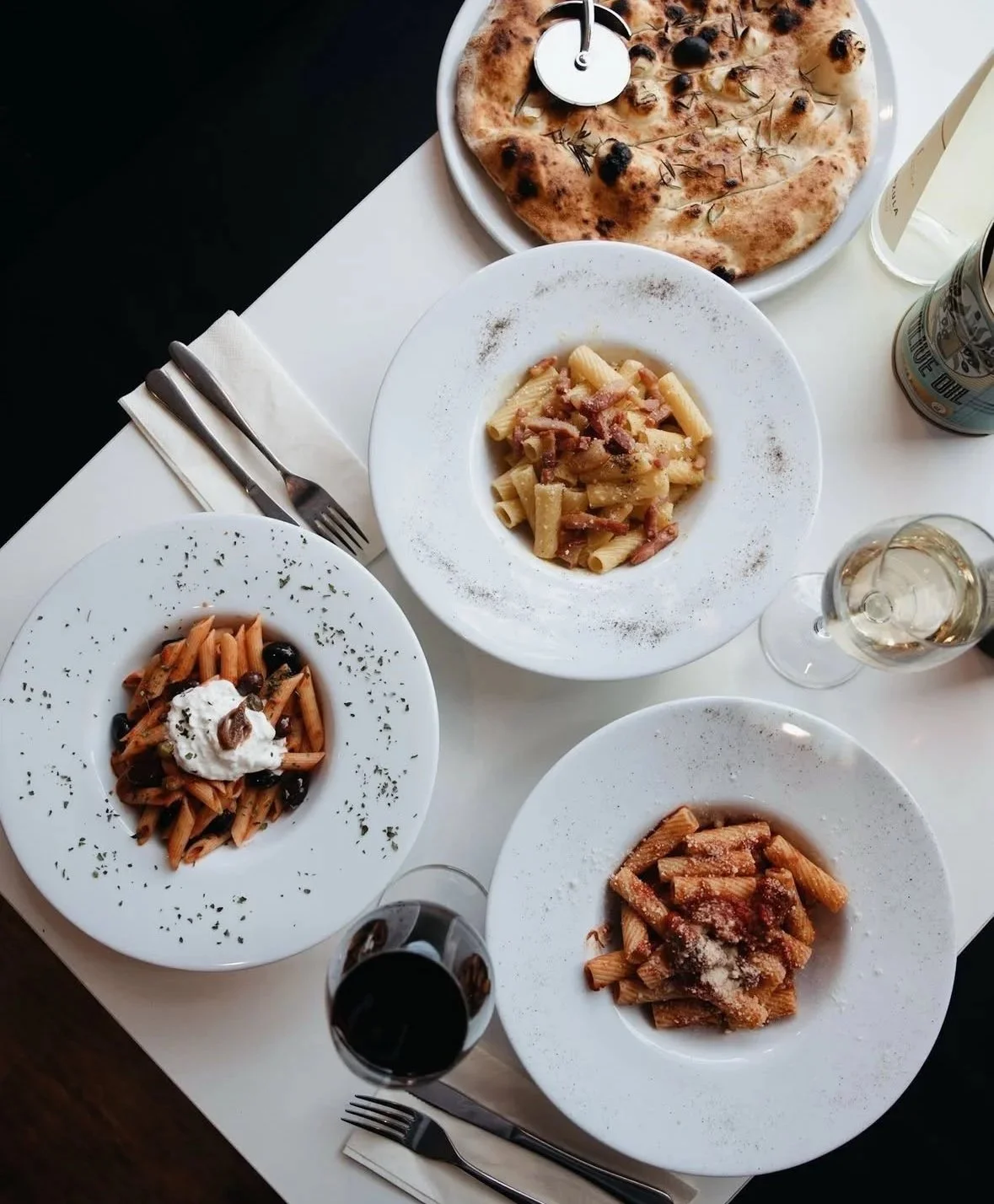 A table with four plates of pasta, a pizza, a glass of white wine, a bottle of water, and cutlery.