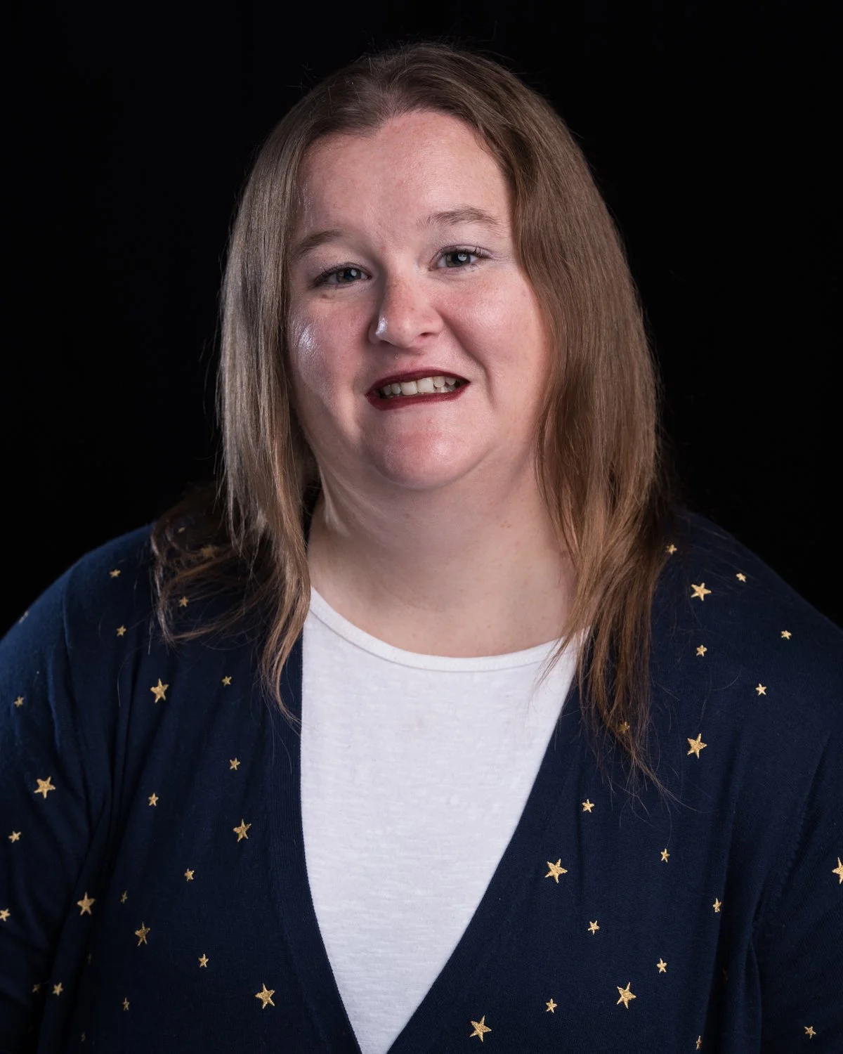 Portrait of a smiling woman with light skin, shoulder-length light brown hair, wearing a white shirt and a dark blue cardigan with gold star patterns, against a black background.