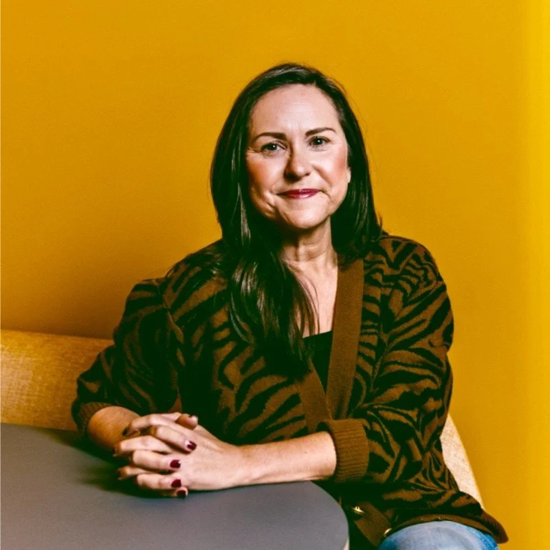 A woman with long dark hair, wearing a brown and black animal print cardigan, sitting at a table against a yellow background, with her hands clasped on the table and a slight smile.