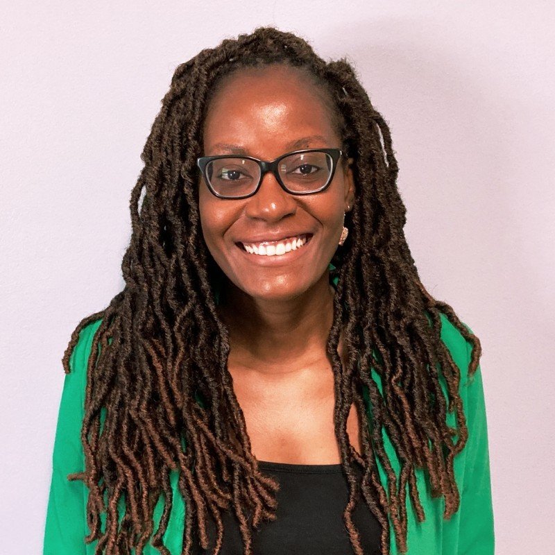A woman with long dreadlocks, wearing glasses, a black top, and a green blazer, smiling at the camera against a plain light background.