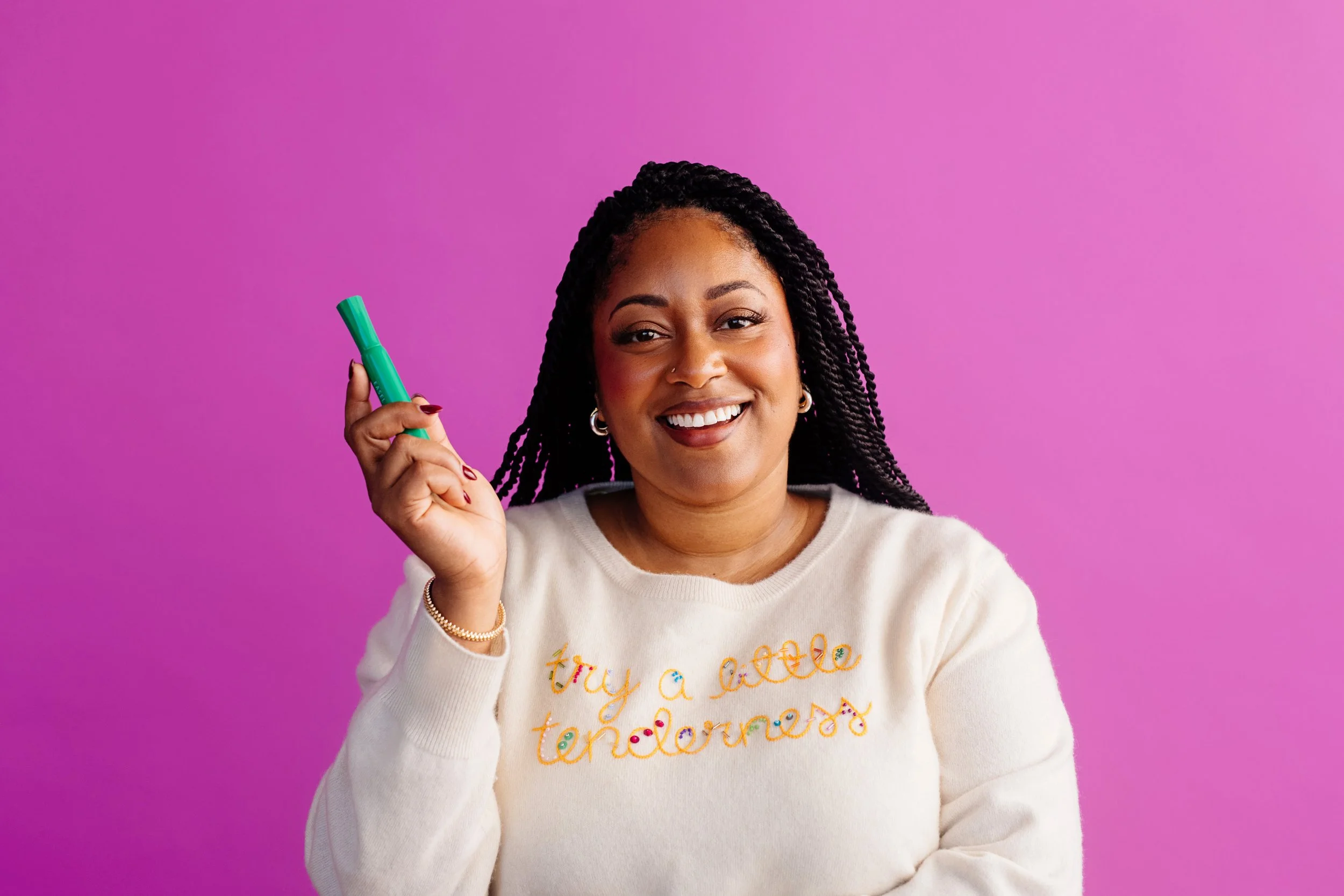 A smiling woman with braided hair wearing a cream-colored sweatshirt with colorful embroidery, holding a green marker against a pink background.