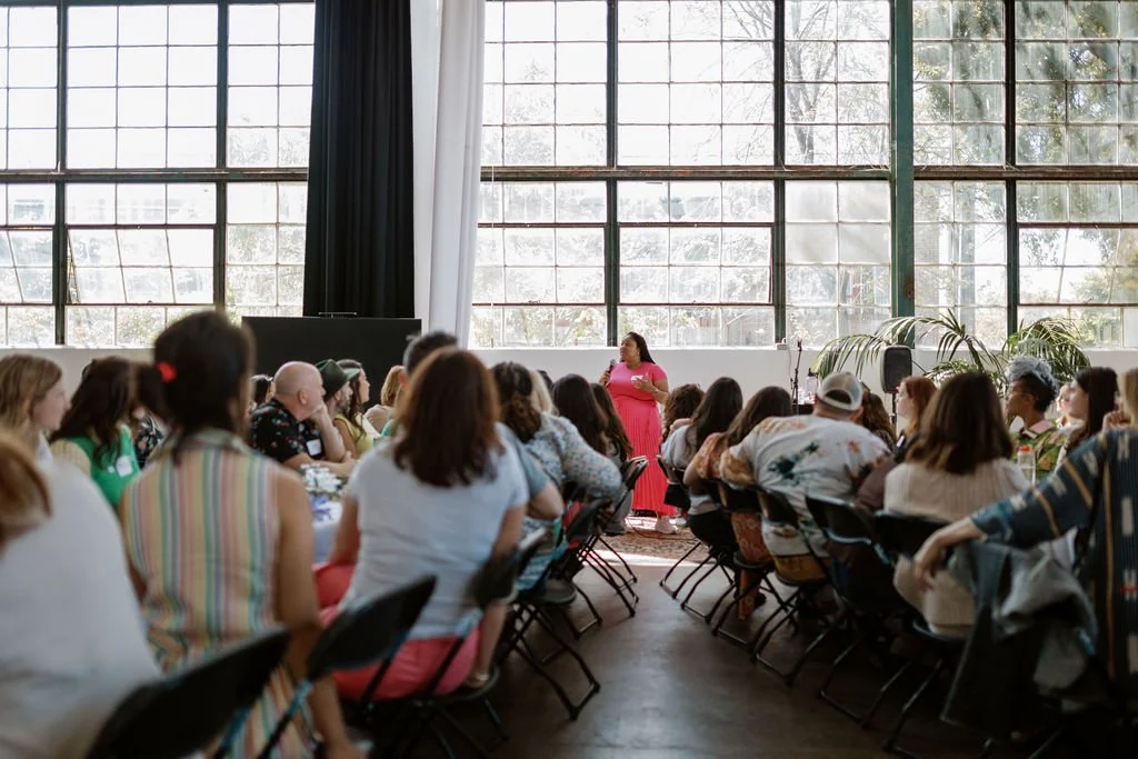 A woman in a pink dress is standing in front of a microphone, speaking to an audience seated at round tables inside a large room with tall, multi-pane windows.