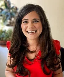 A woman with long dark hair smiling, wearing a red dress, in front of a decorated Christmas tree.