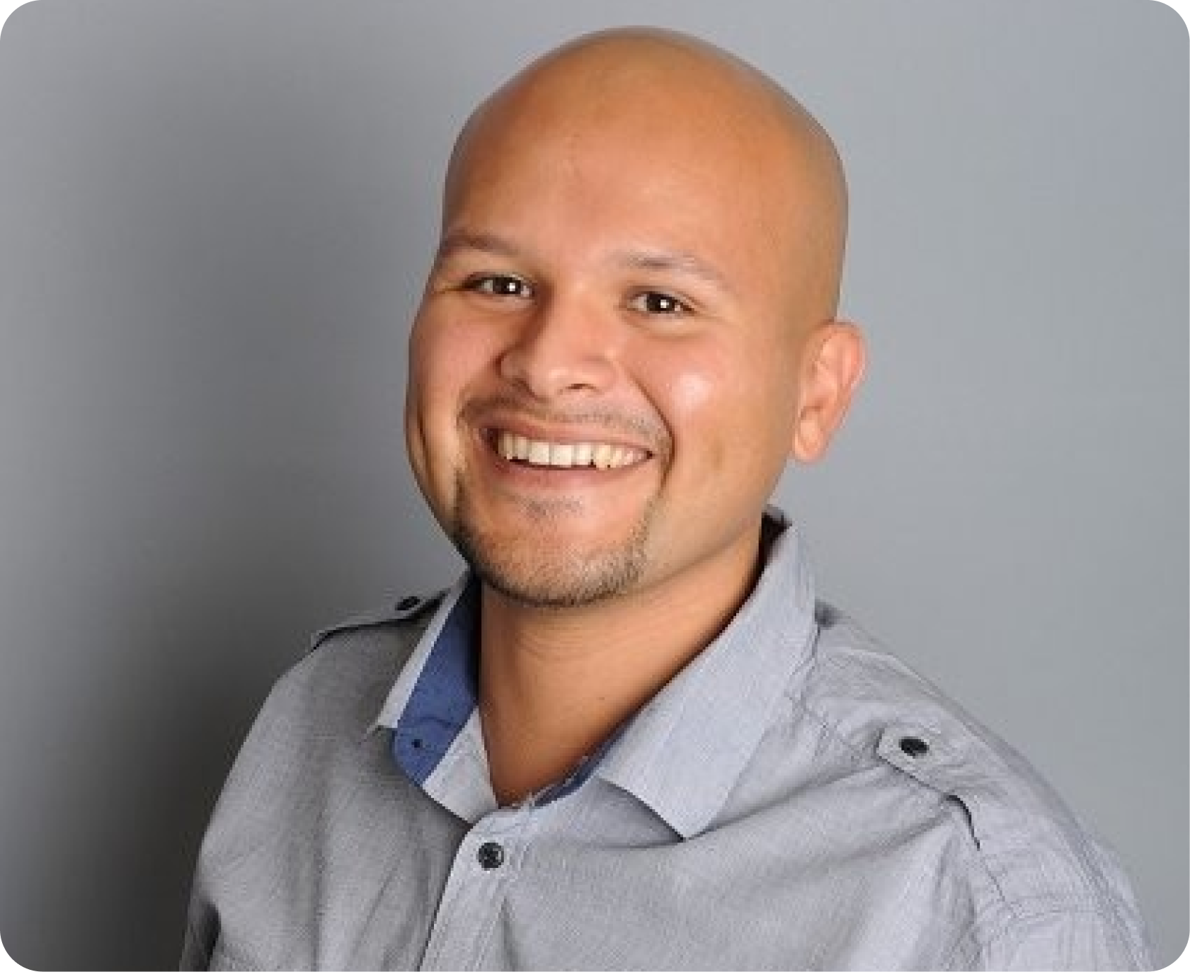 A smiling man with a shaved head and goatee, wearing a light gray collared shirt with a darker blue inner shirt, posed against a gray background.