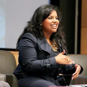 Woman with curly black hair, wearing a dark blazer, smiling, holding a smartphone in a professional setting.