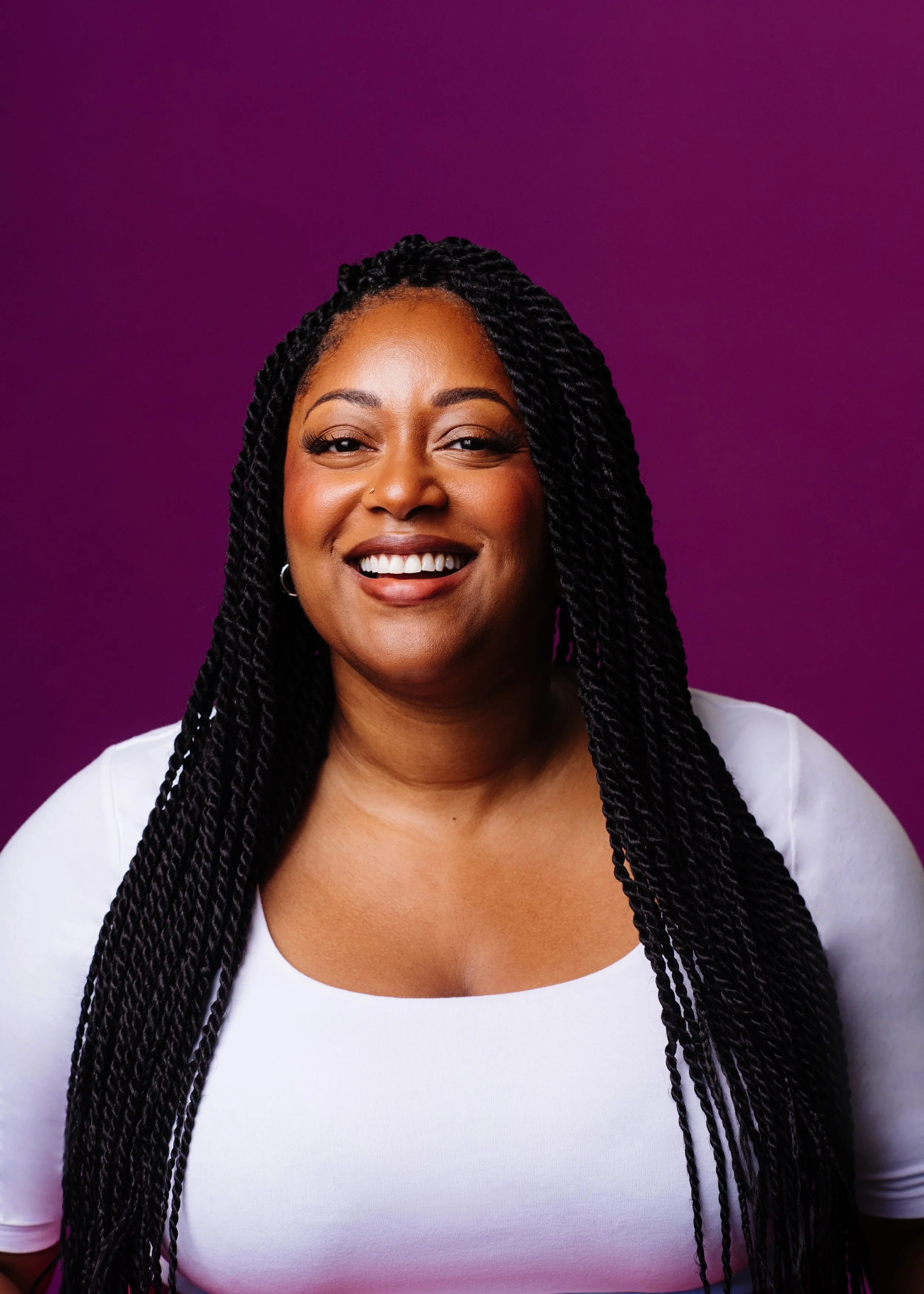 Close-up portrait of a smiling woman with long braided hair, wearing a white top, against a purple background.