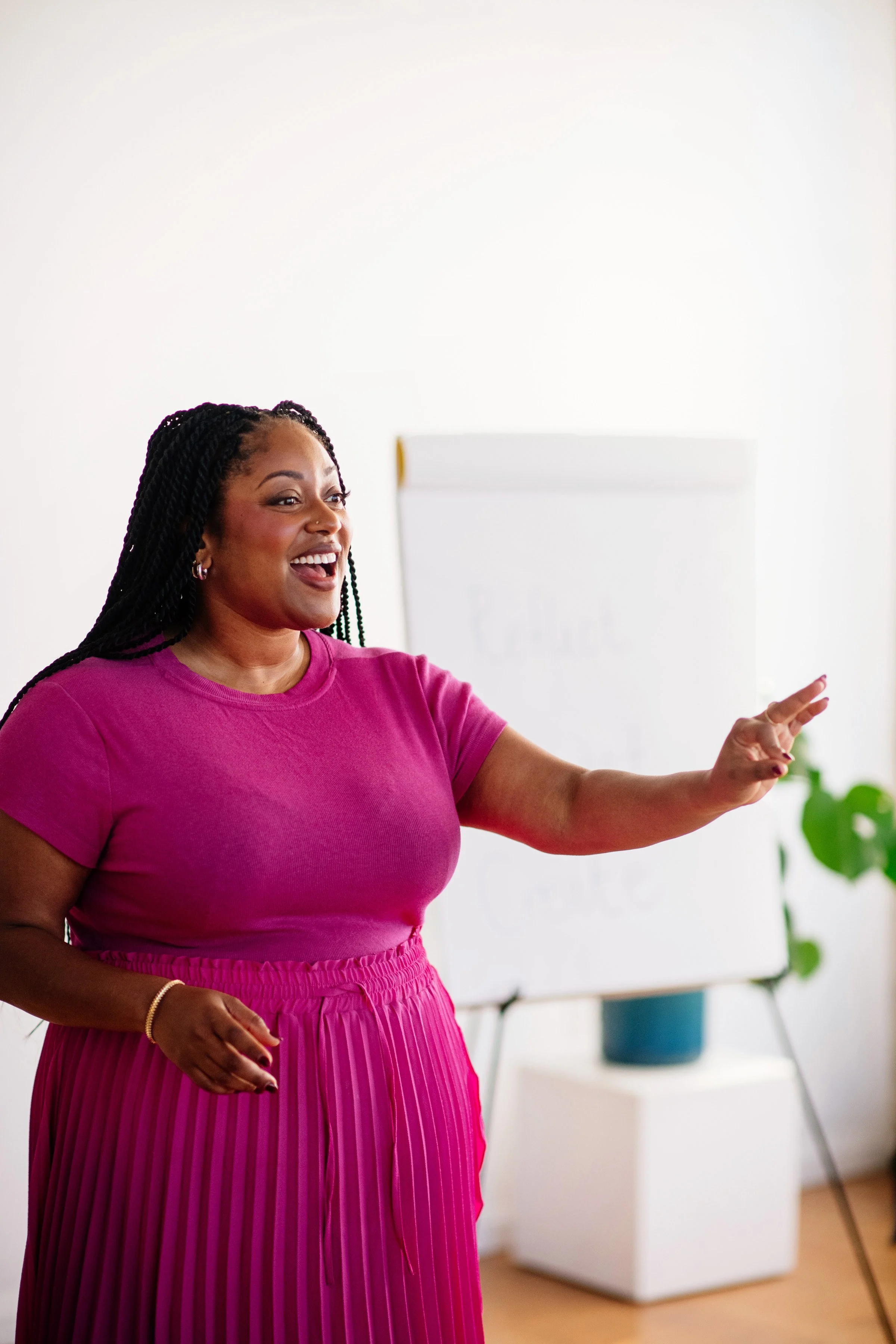 A woman with braided hair, wearing a pink top and pink pleated skirt, smiling and gesturing with her right hand during a presentation in a bright room with a whiteboard and a potted plant.