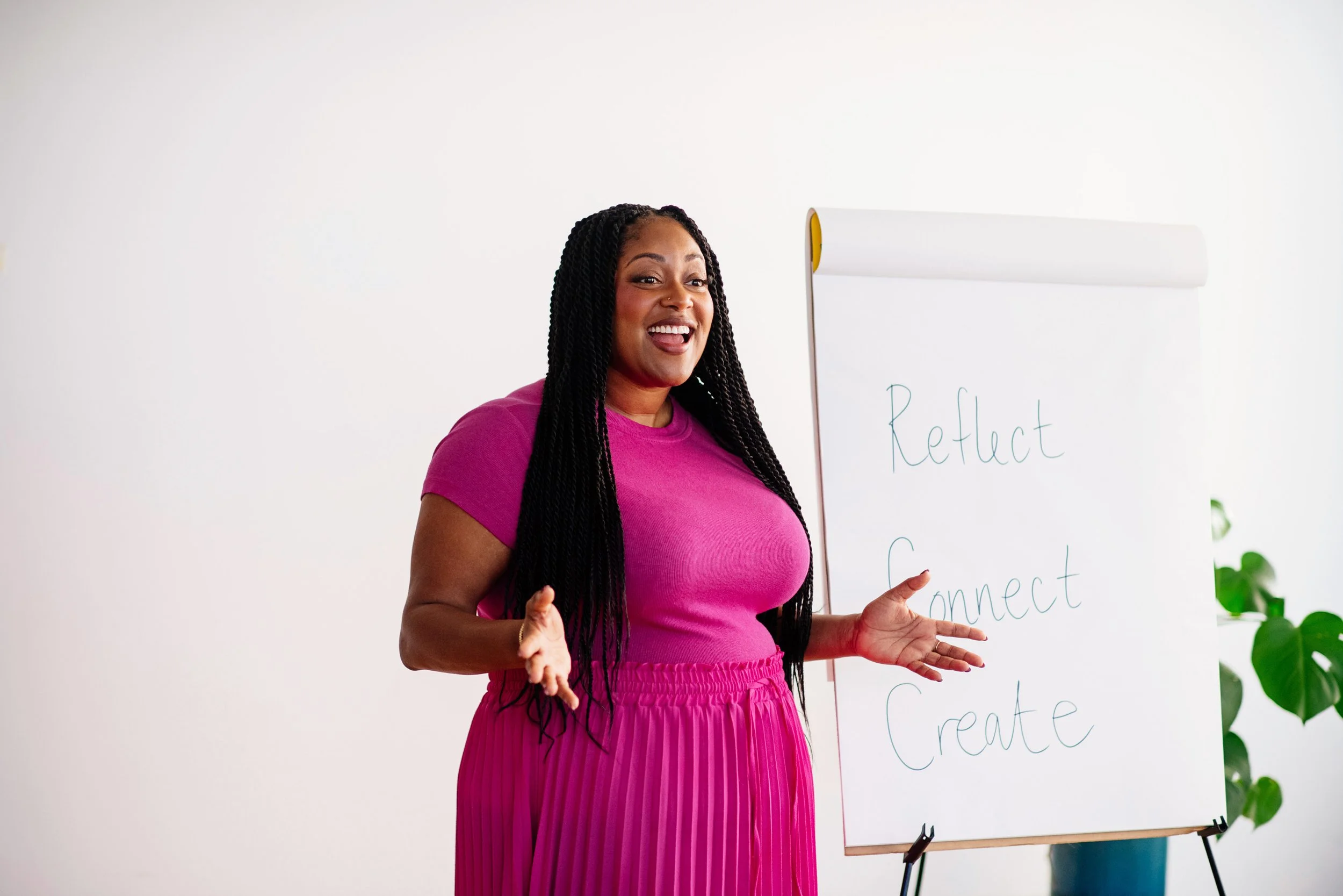 A woman with long braided hair, wearing a pink top and matching skirt, smiling and gesturing with her hands as she stands next to a whiteboard with words like 'Reflect' and 'Connect' written on it.