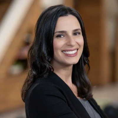 A woman with dark hair smiling, wearing a black blazer, in an indoor setting with wooden details in the background.